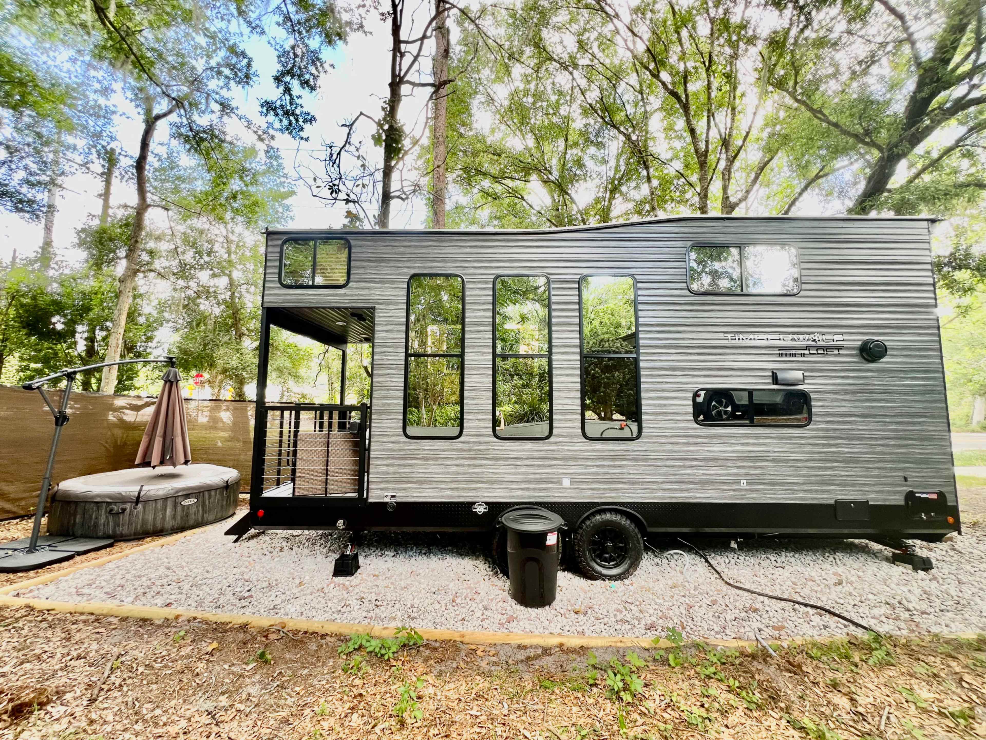 A modern trailer with large windows is parked beside a round hot tub in a wooded area.