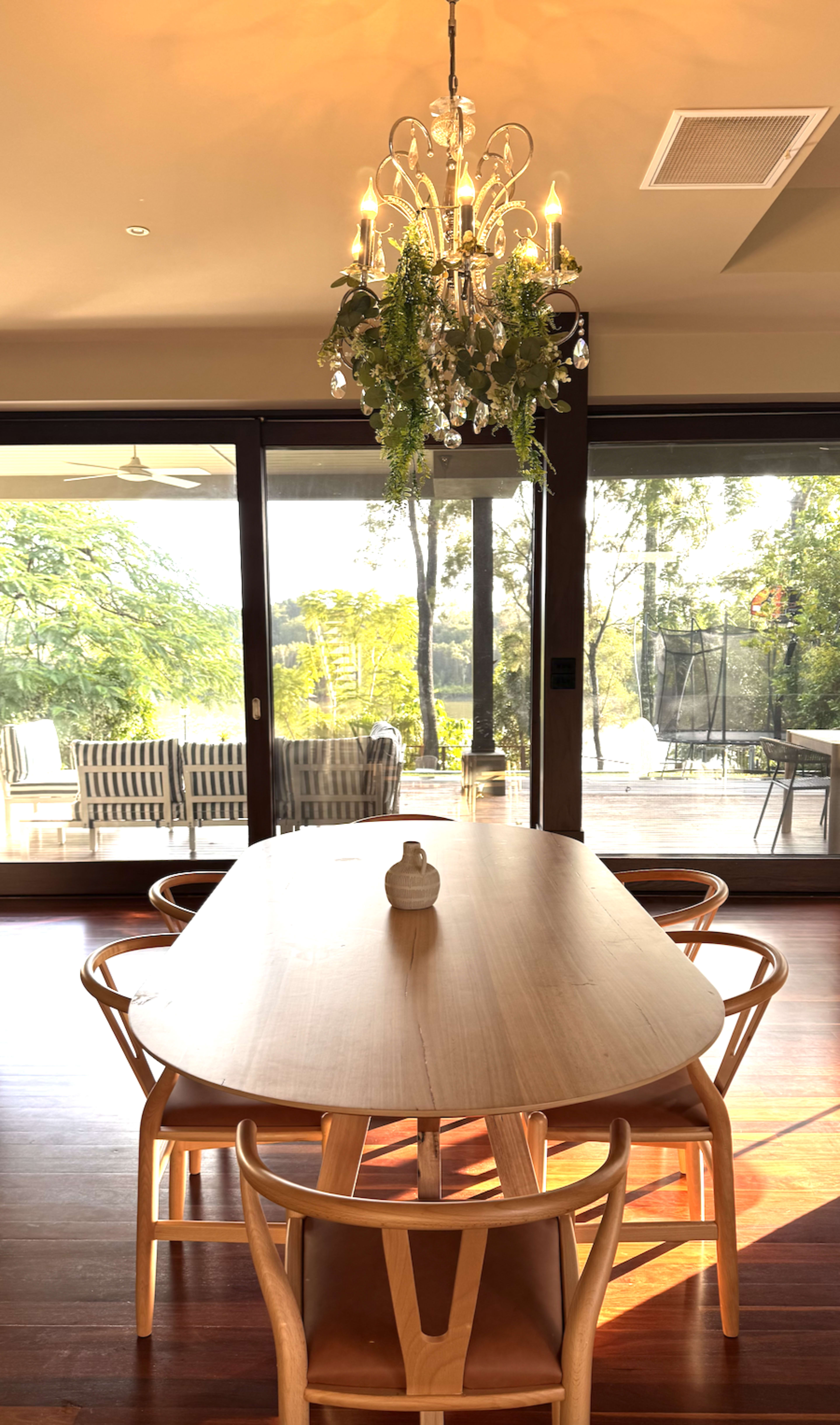 A wooden dining table with curved chairs is set beneath a chandelier in a room with large glass doors that open to a view of greenery outside.