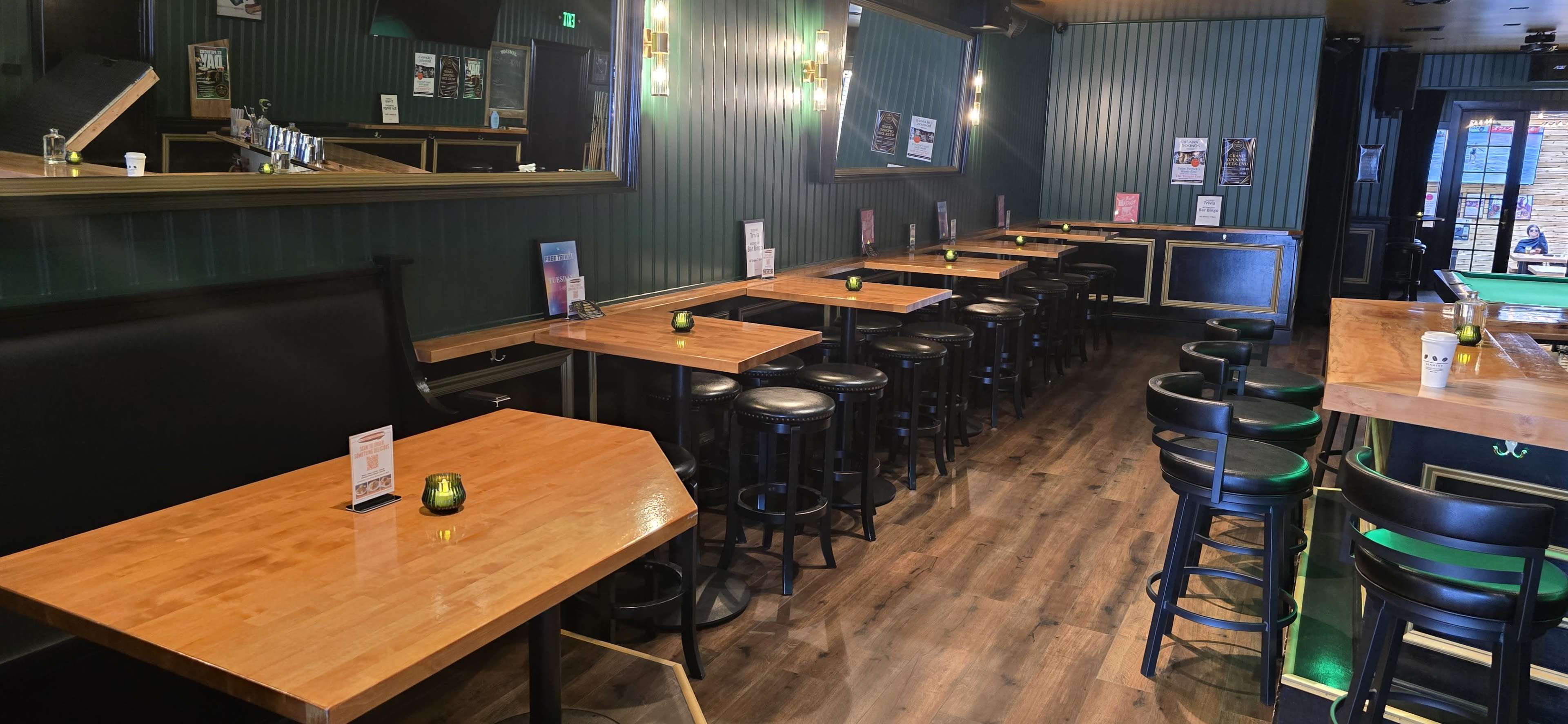 The image shows a brightly lit bar with wooden tables and high stools arranged in parallel lines against green walls.