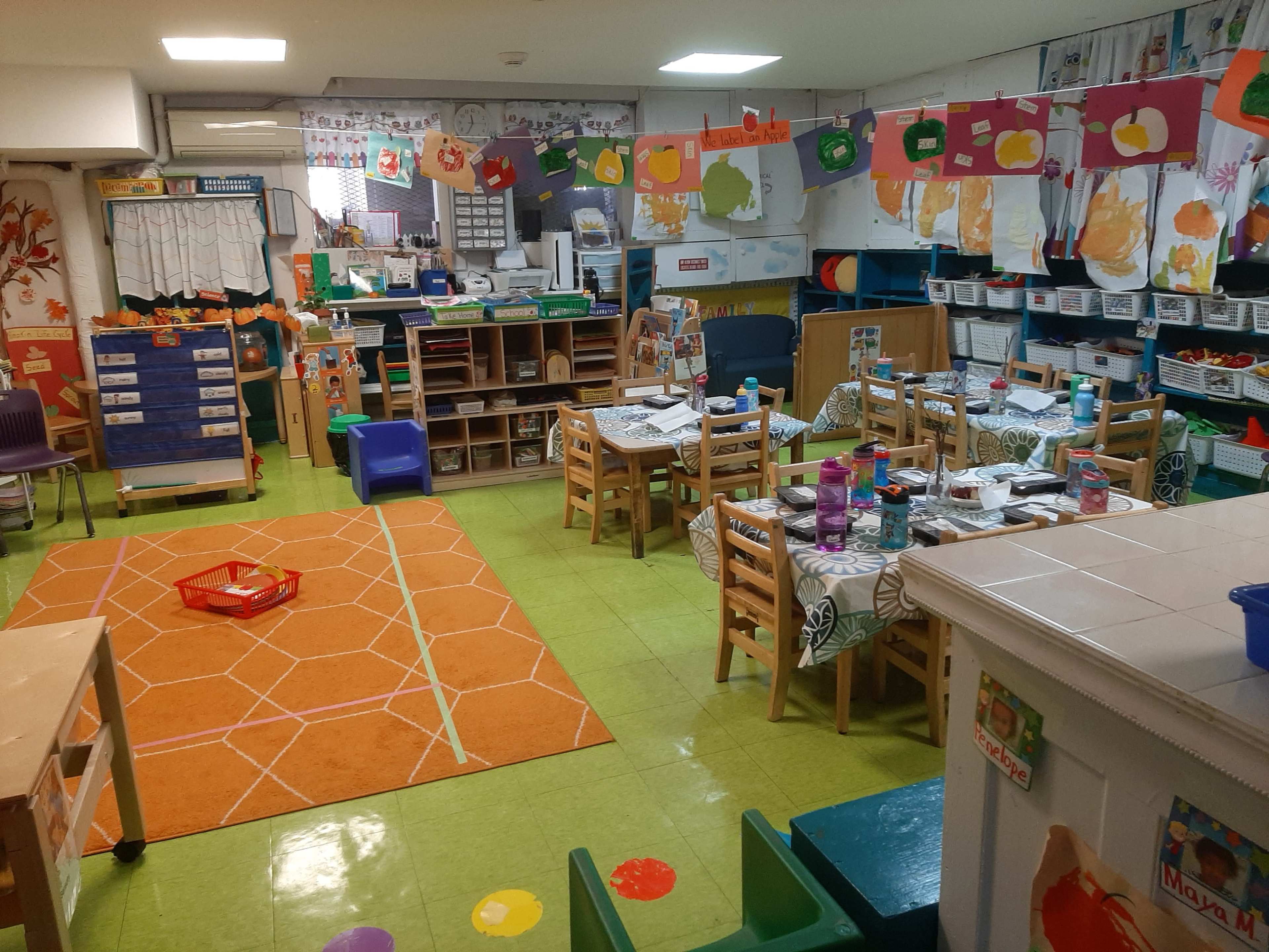 A colorful classroom features tables with chairs, artwork on the walls, and various educational materials organized on shelves.