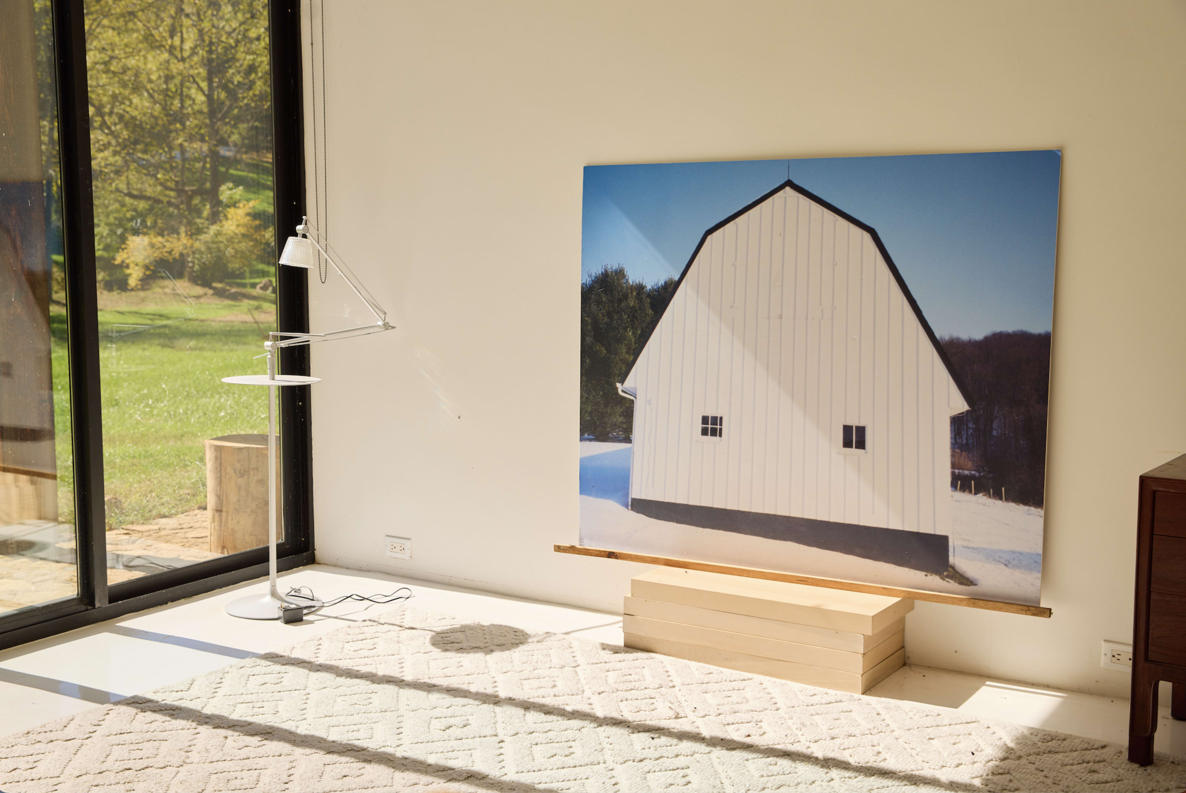 A large photo of a white barn is displayed on a stand in a room with a glass wall, overlooking a green landscape.