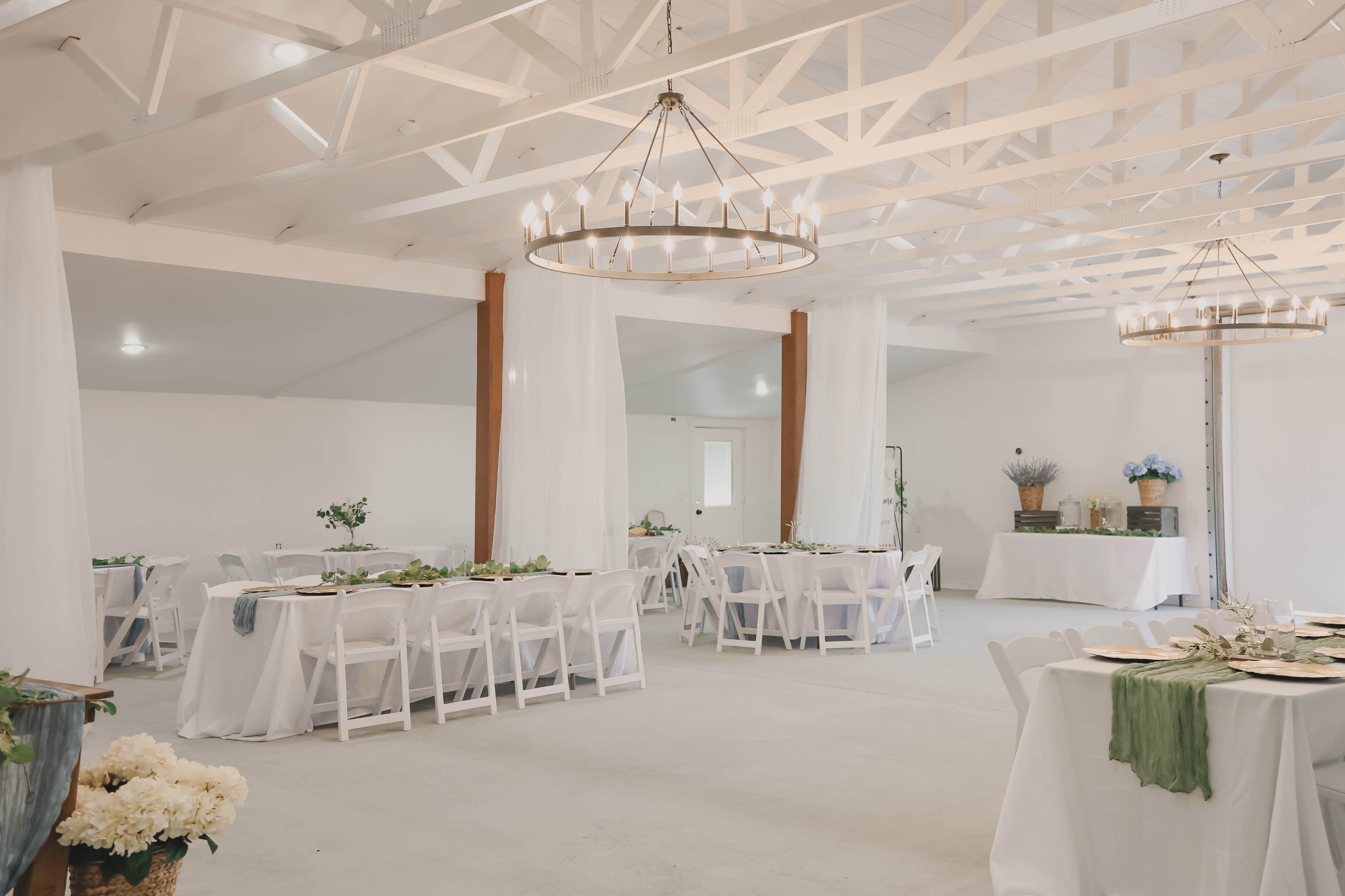 The image shows a spacious, elegantly arranged dining area with round tables set for a meal, white drapery, and large chandeliers hanging from a white beamed ceiling.