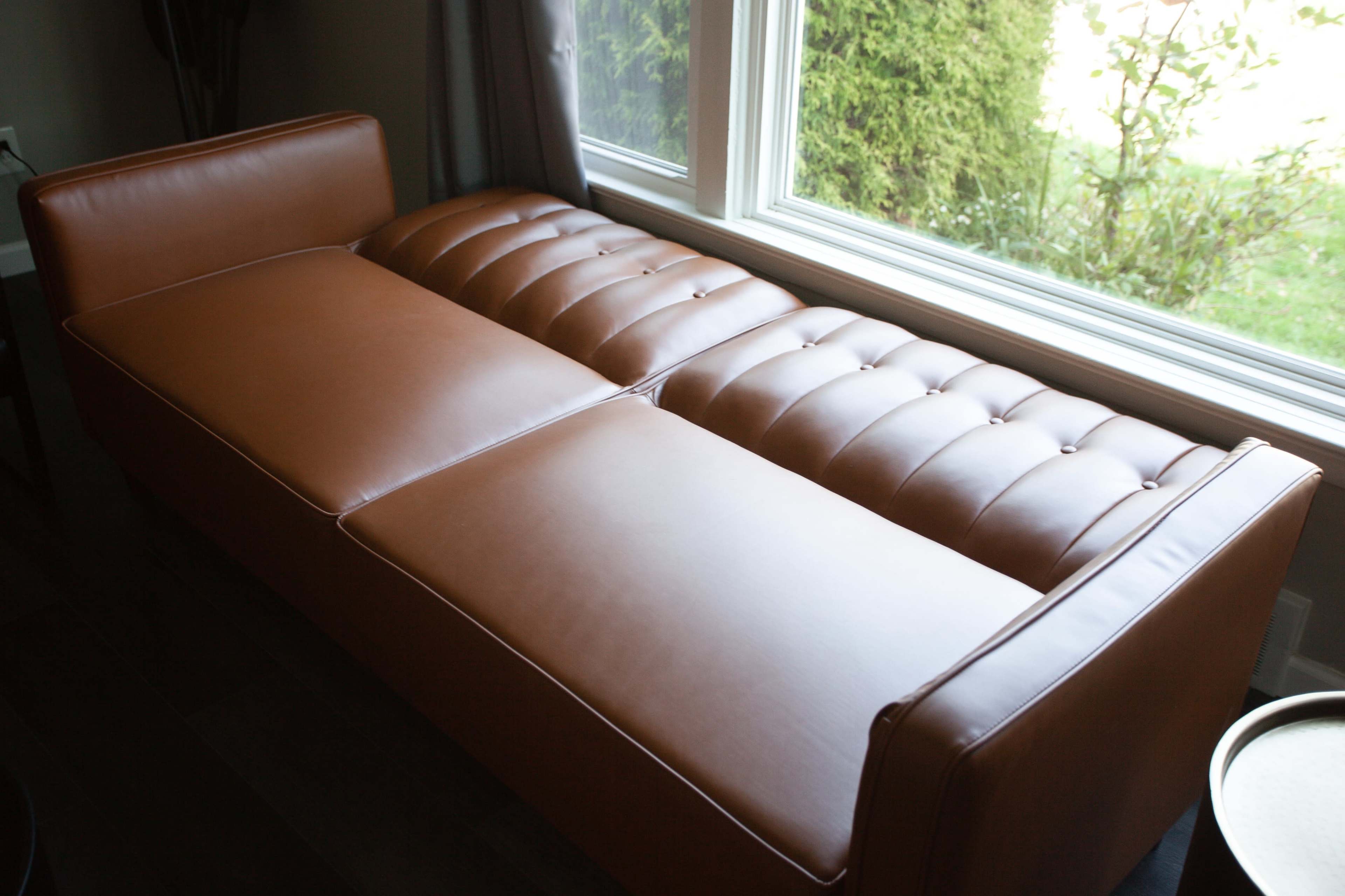 A brown leather sofa with a tufted design is positioned next to a window.