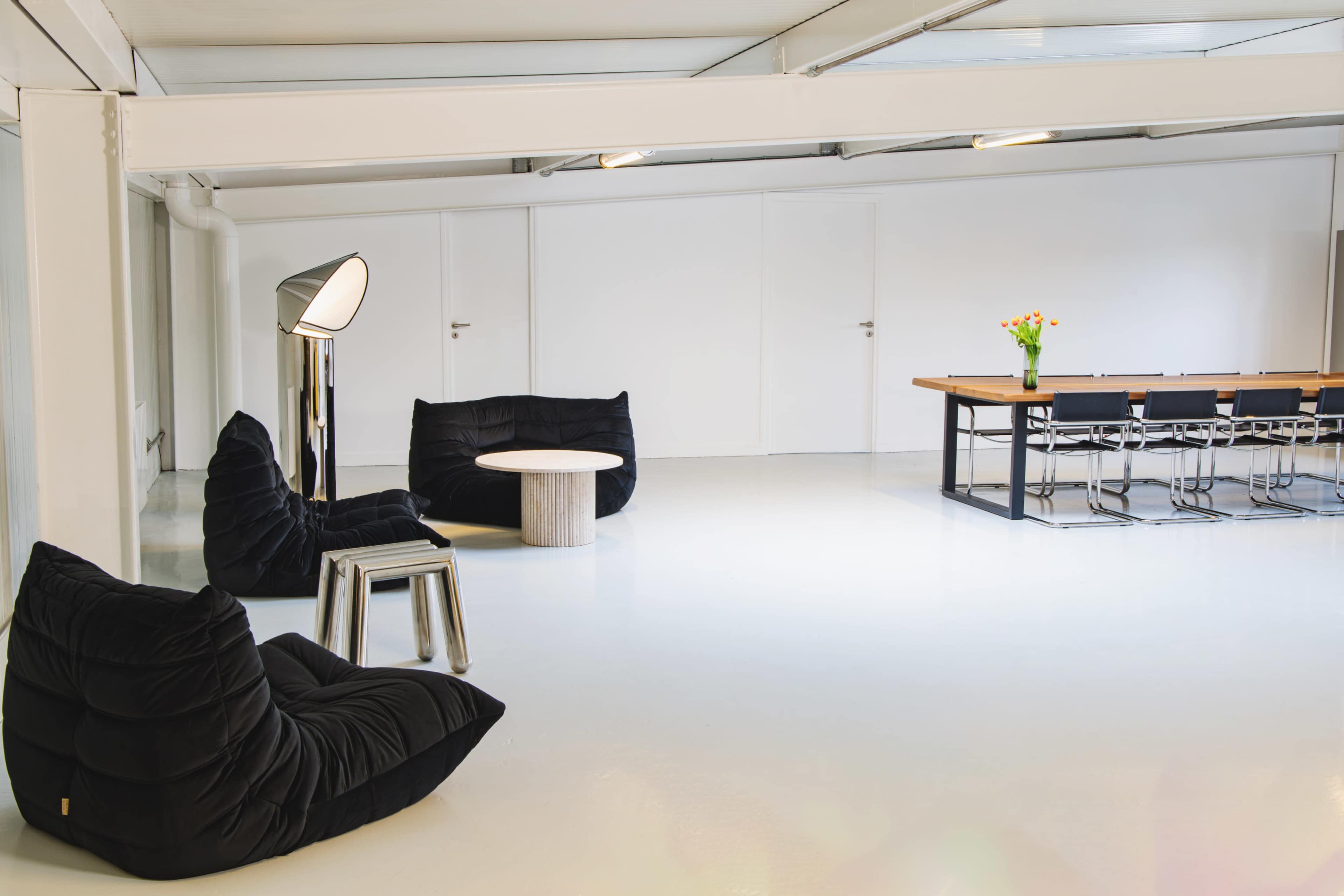 The scene features a minimalist room with black beanbag chairs, a small round table, a floor lamp, and a long dining table with chairs, all set against a white backdrop.