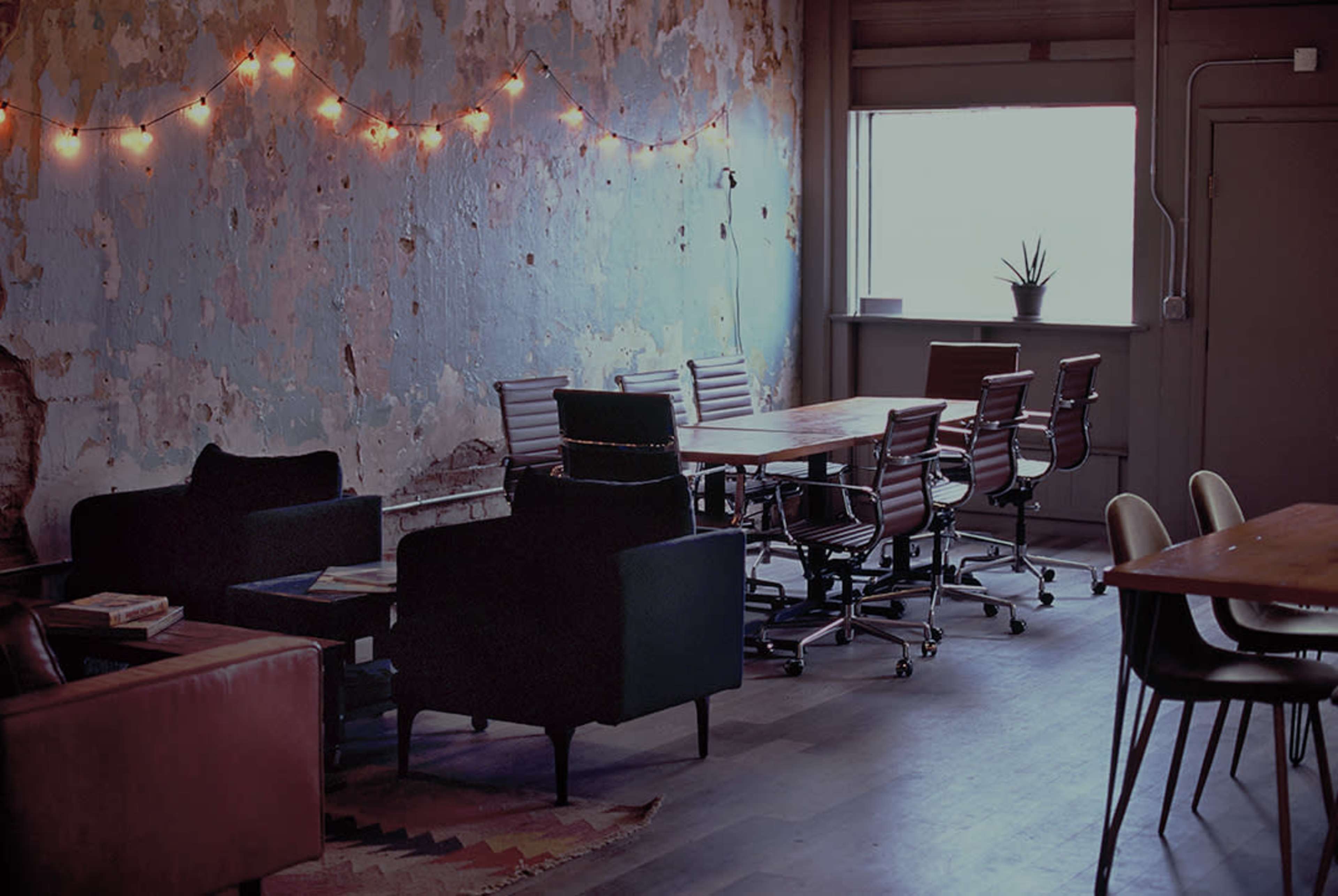 A modern workspace features wooden tables, rolling chairs, and a textured blue wall adorned with string lights.