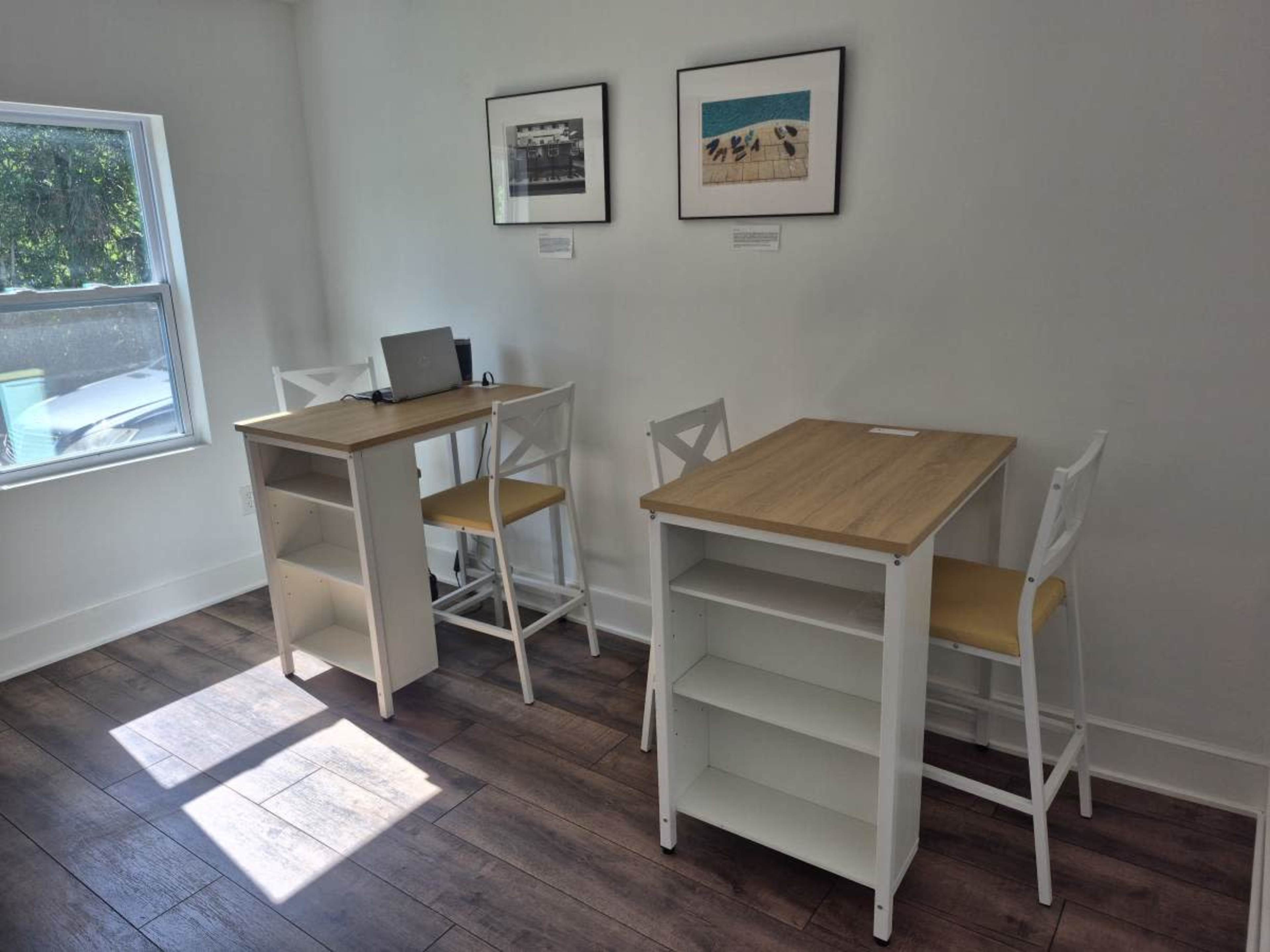 The image shows a bright room with two small tables and chairs placed against the walls, featuring framed photos on the white walls.