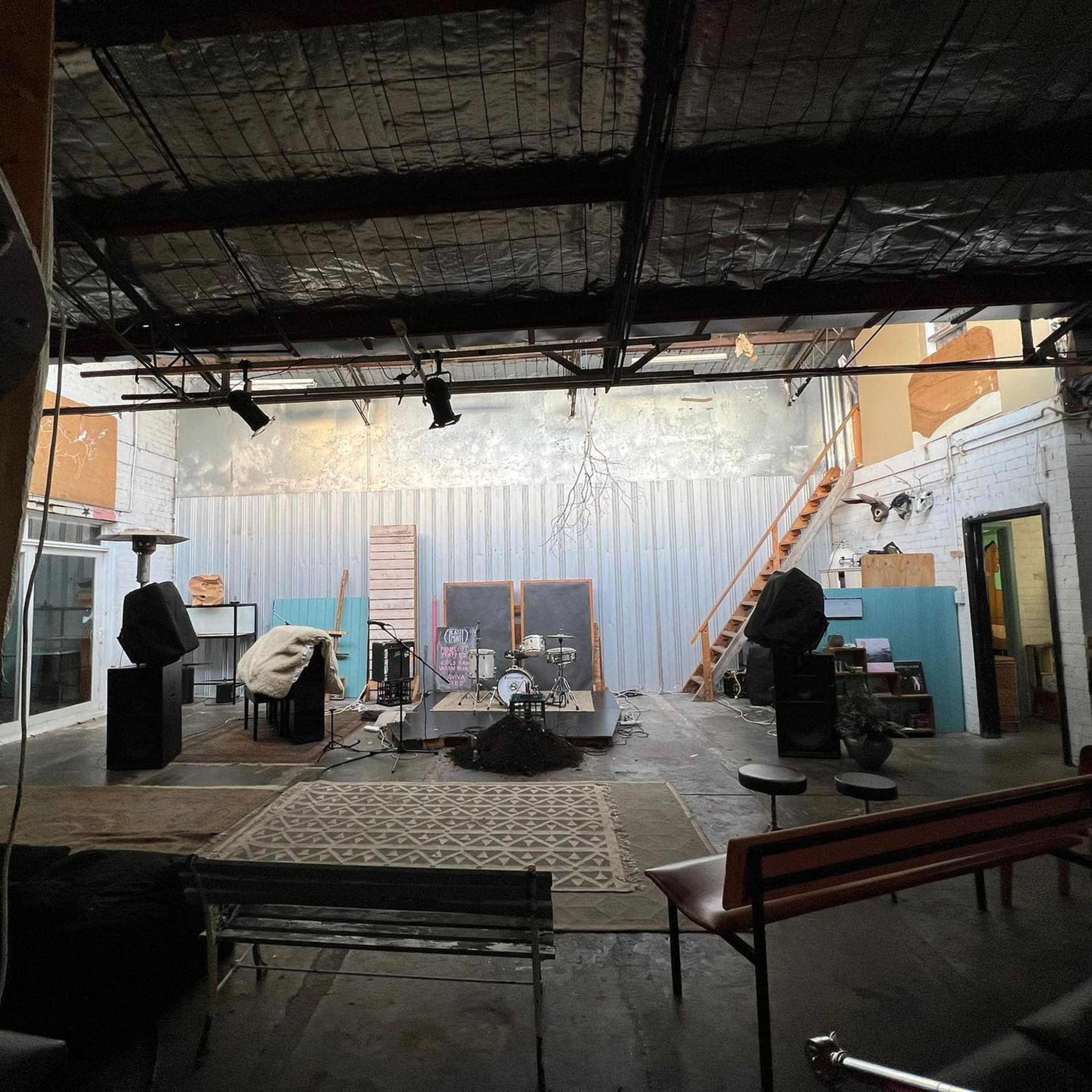 A spacious, empty indoor area with a wooden staircase, scattered musical equipment, and a few chairs arranged on a textured floor.