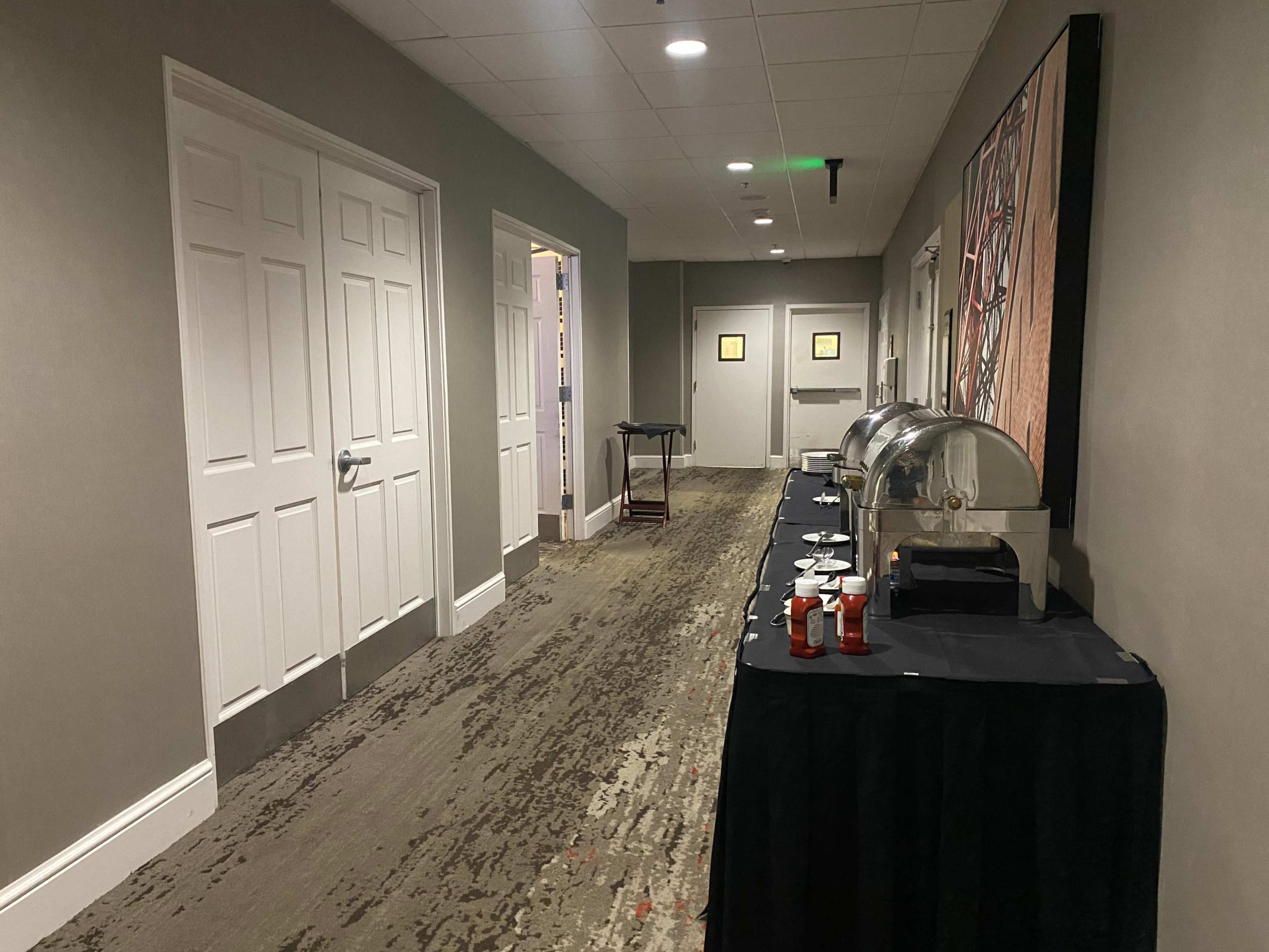 The image shows a hallway with two double doors, a cart, and a table covered in a black cloth holding food warmers and cups.