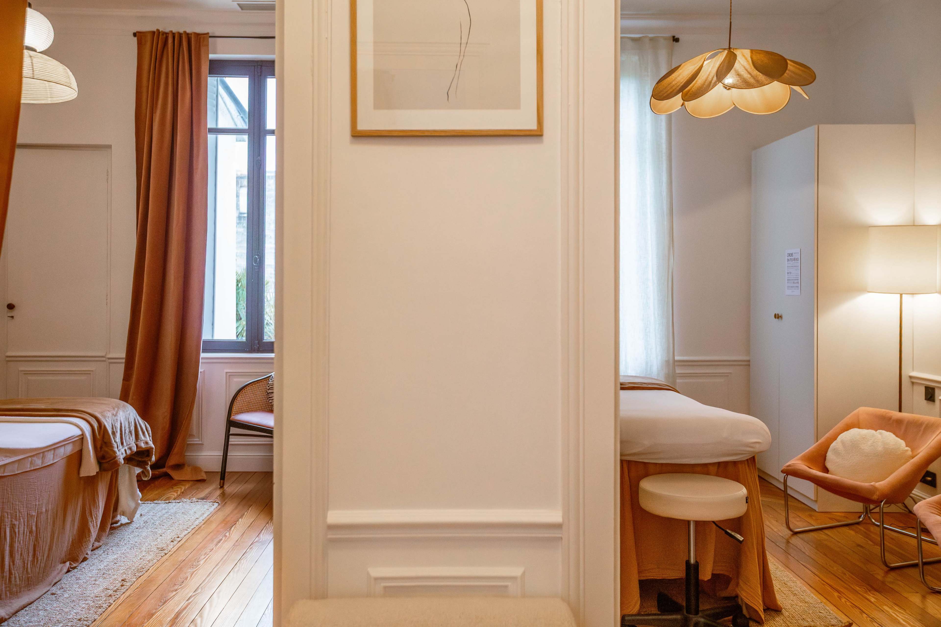 The image depicts a bright and airy interior of a room with two massage tables, wooden flooring, and large windows dressed with curtains.