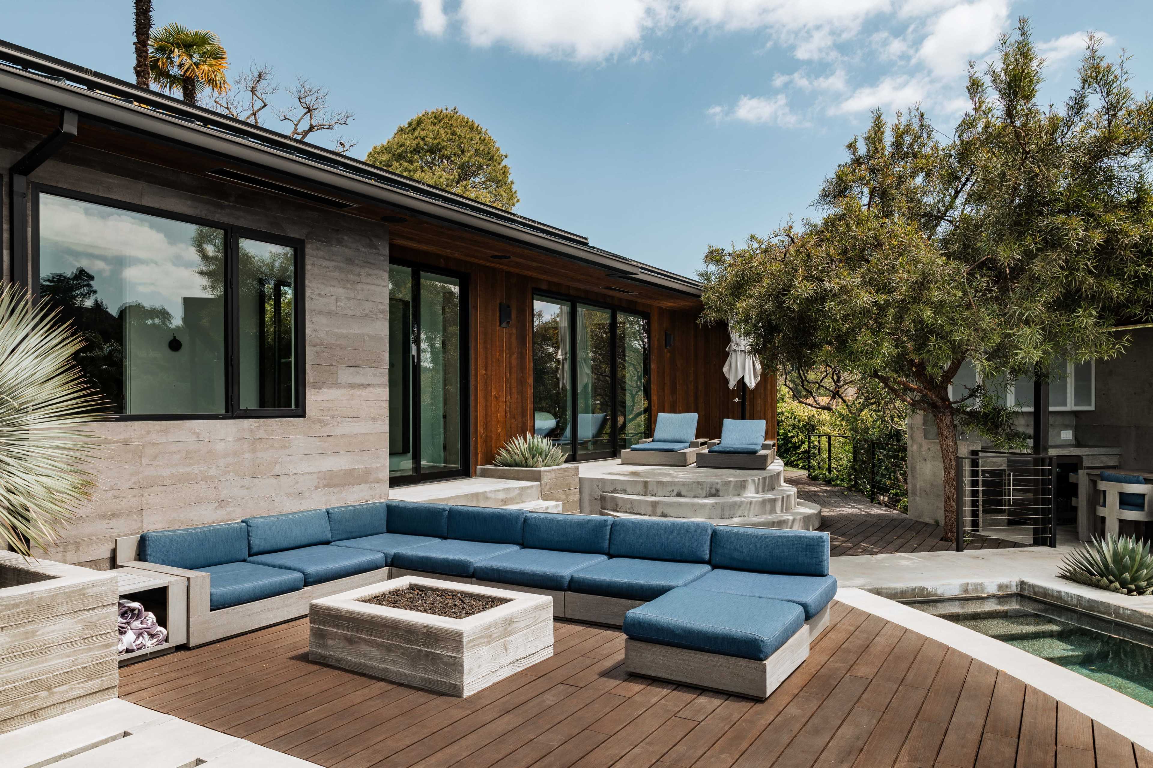 The image shows a modern outdoor patio area featuring a large sectional sofa, a fire pit, and a swimming pool, surrounded by greenery and architectural elements.