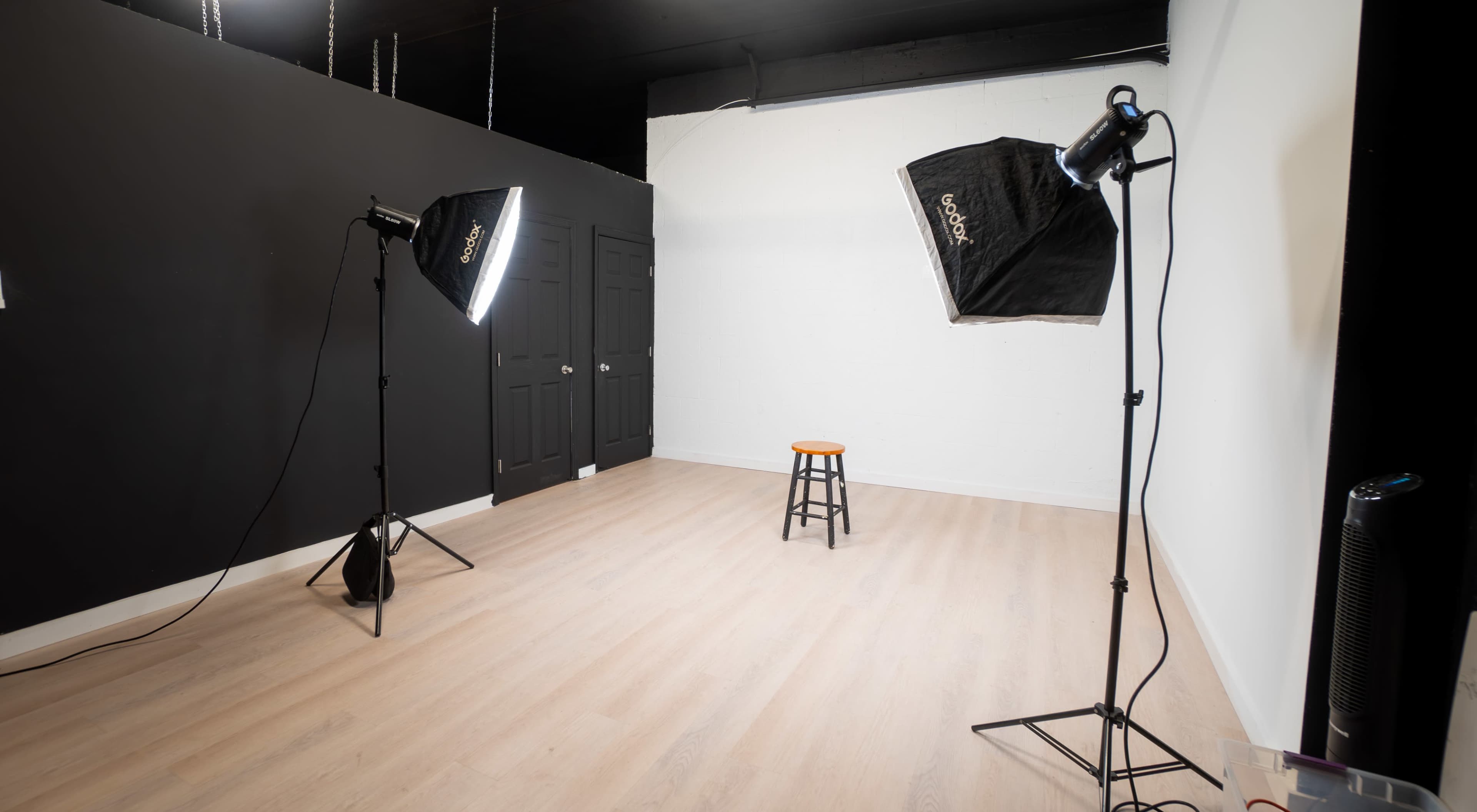 The image shows a photography studio with a black and white backdrop, two softbox lights, and a wooden stool in the center.