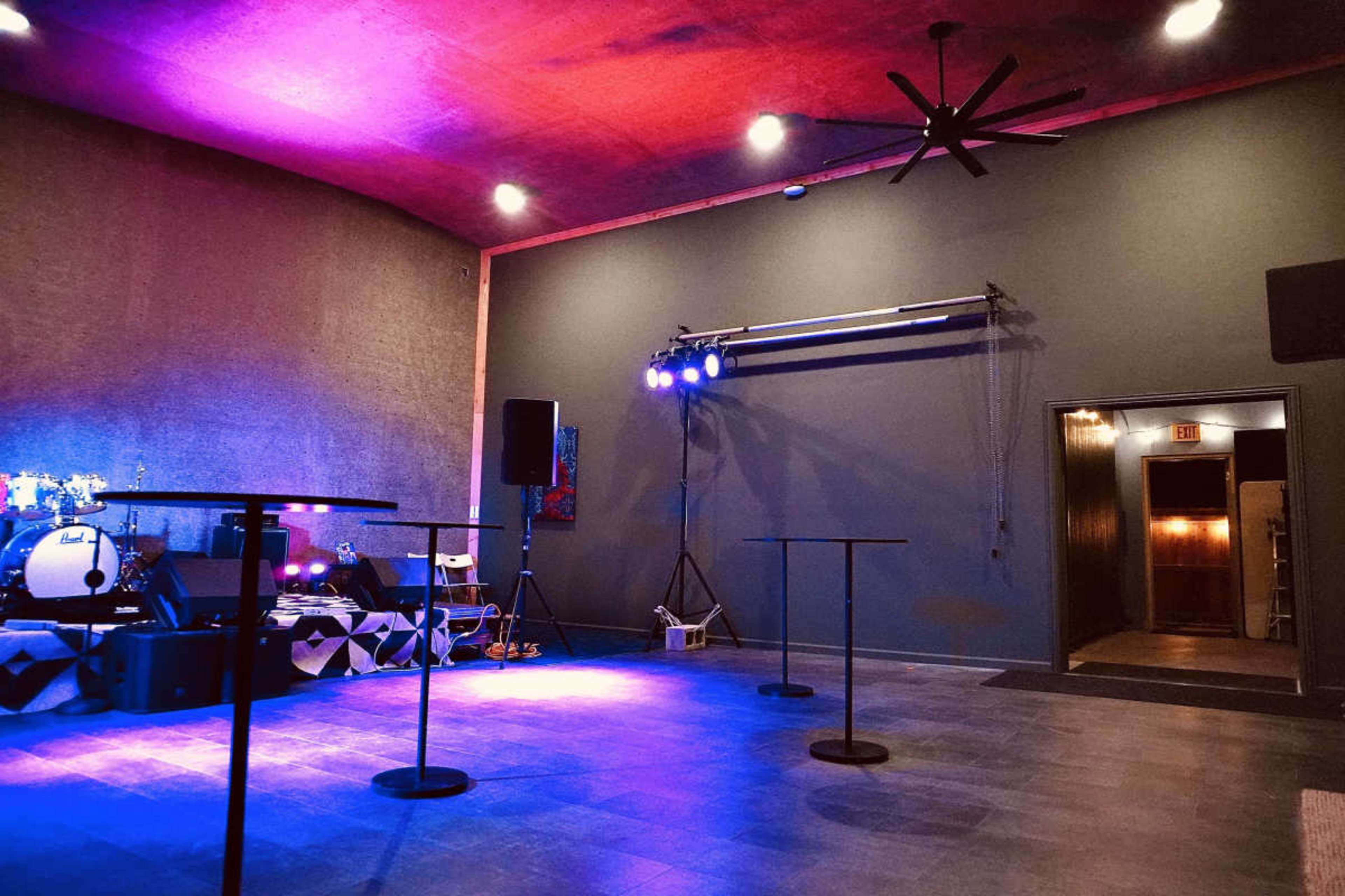 The image shows a dimly lit music venue with round tables, a drum set, and colored stage lights illuminating the space.