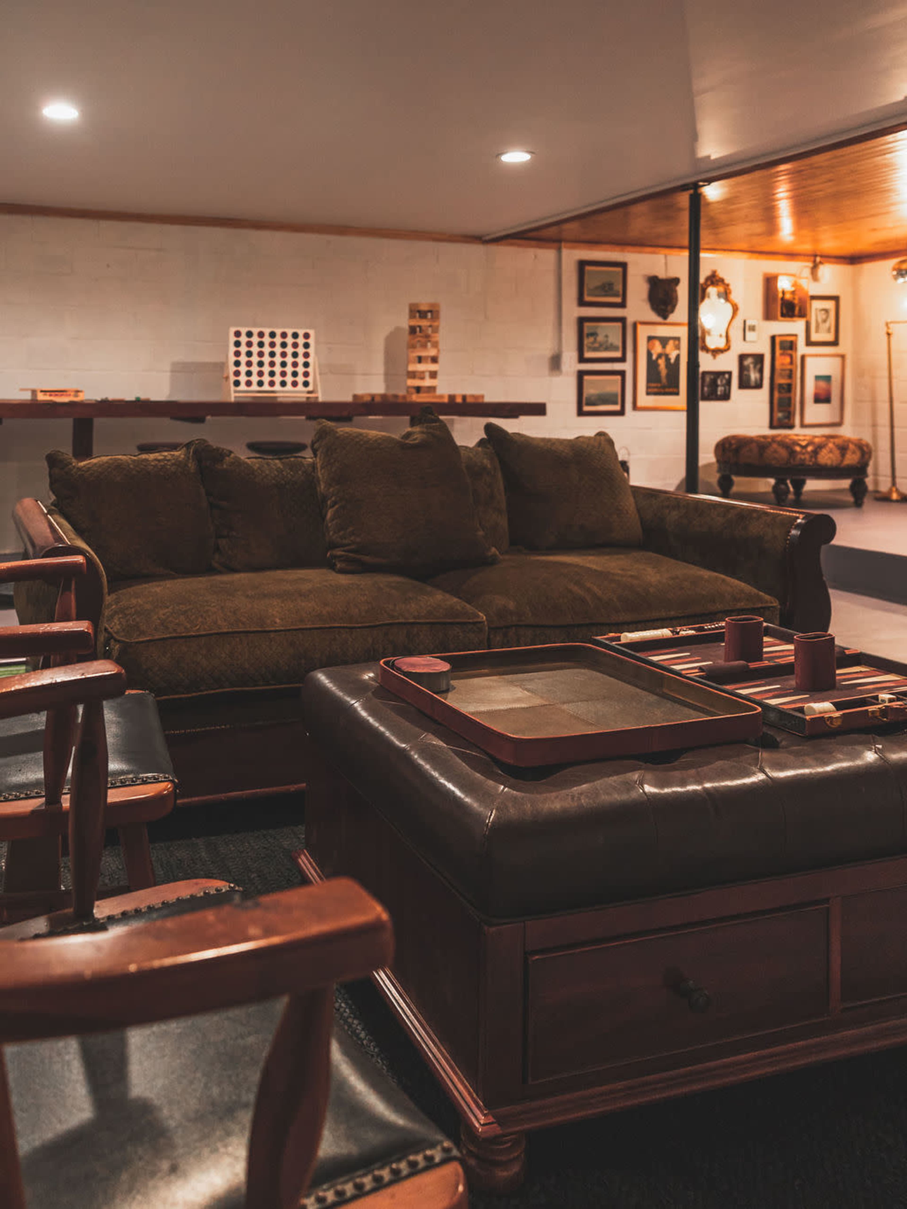 The image shows a cozy living room with a large brown sofa, wooden chairs, and a coffee table featuring a backgammon set, surrounded by framed pictures on the walls.