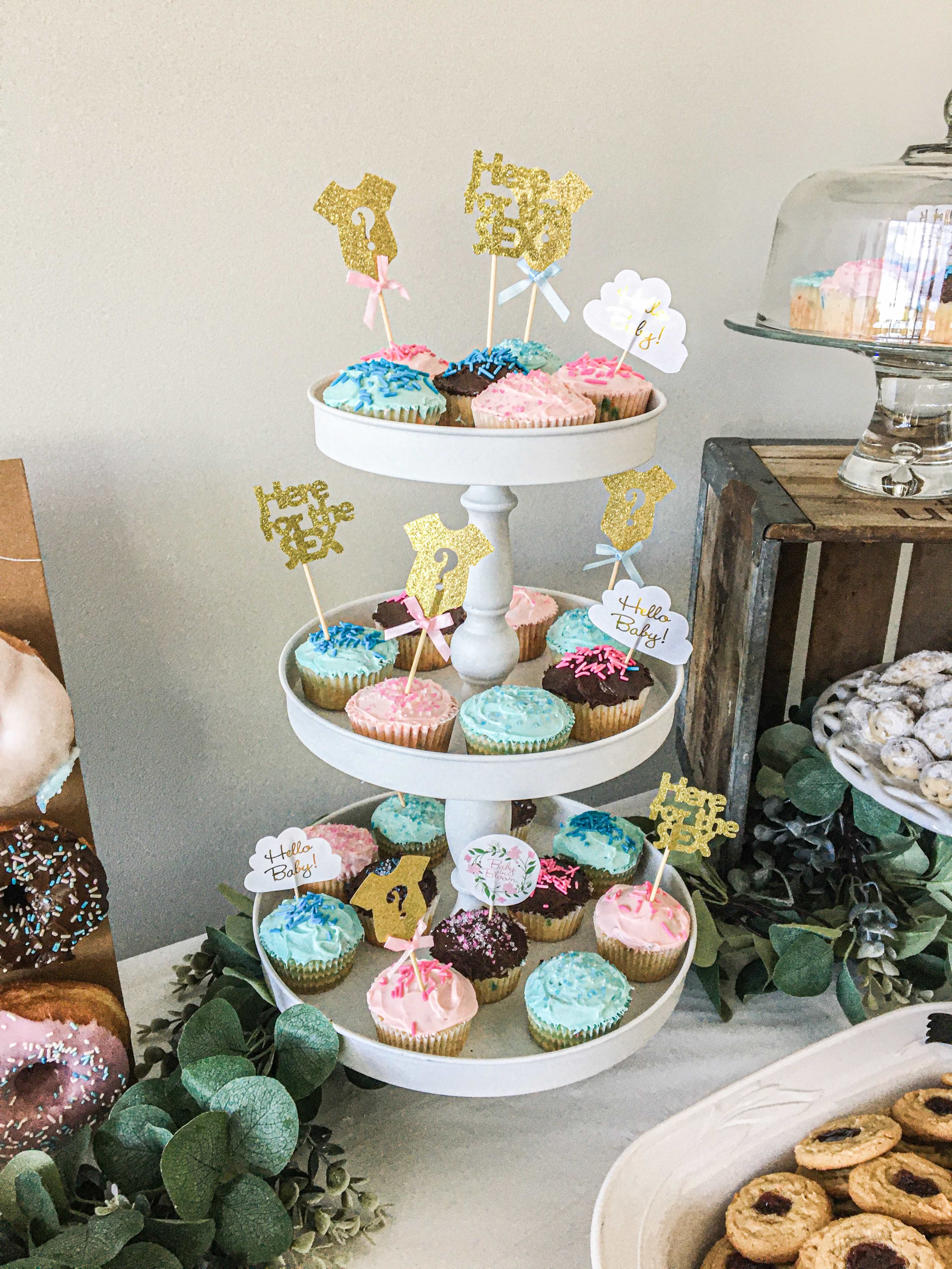 A three-tiered stand filled with colorful cupcakes decorated with various toppers, surrounded by a rustic display of donuts and greenery.