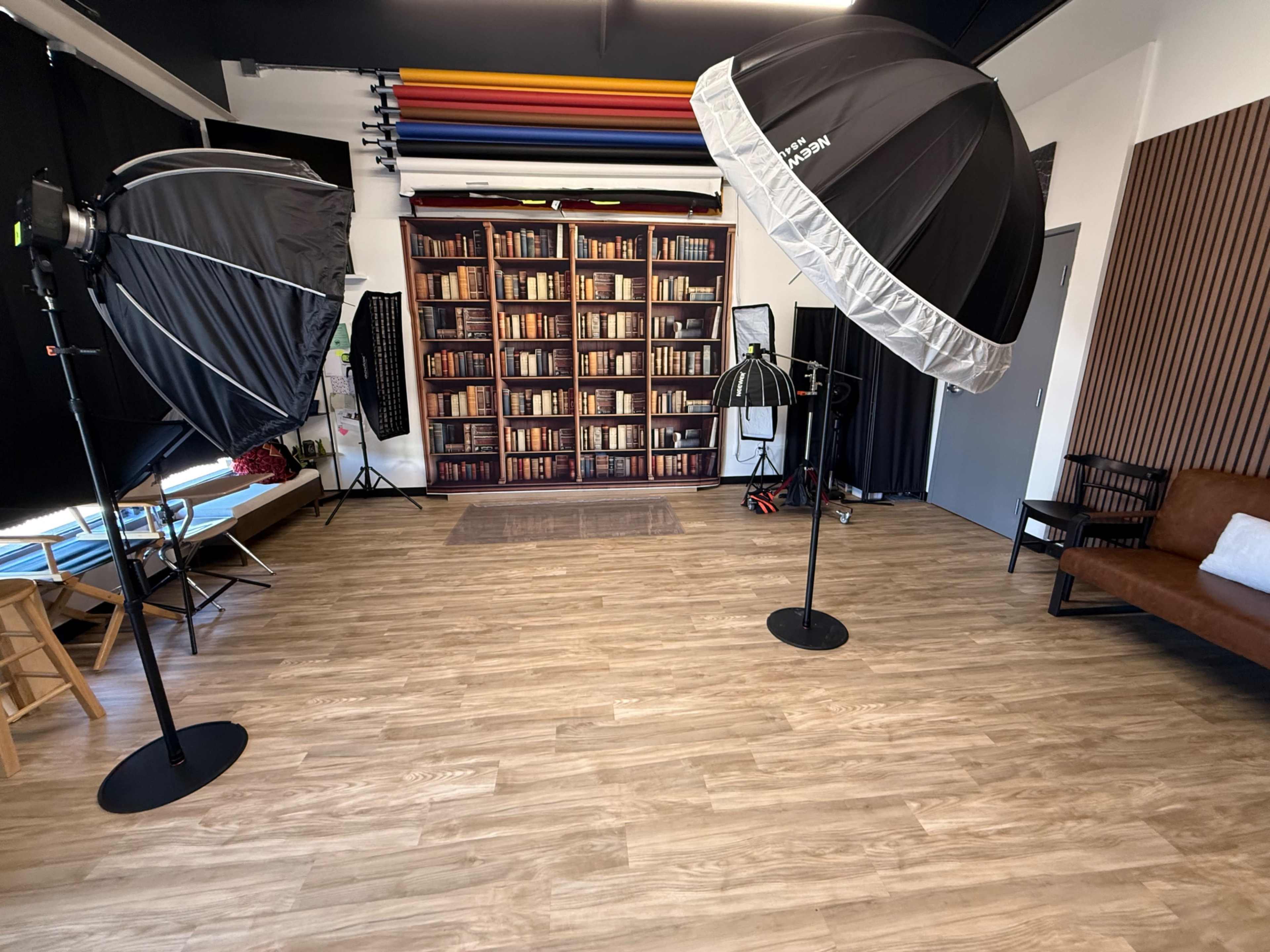The image shows a photography studio with backdrop shelves filled with books, two large softbox lights, and a brown sofa.