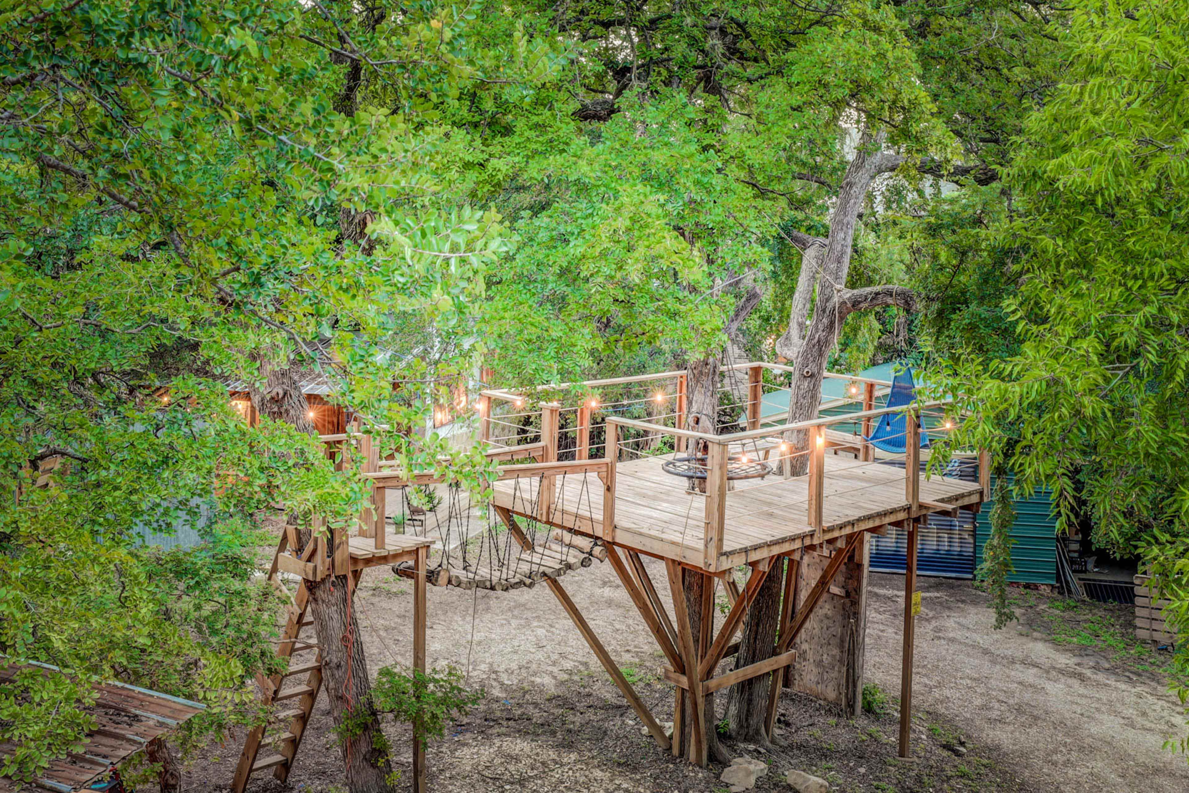 A wooden treehouse platform is elevated among green trees, connected by a rope bridge to another structure.