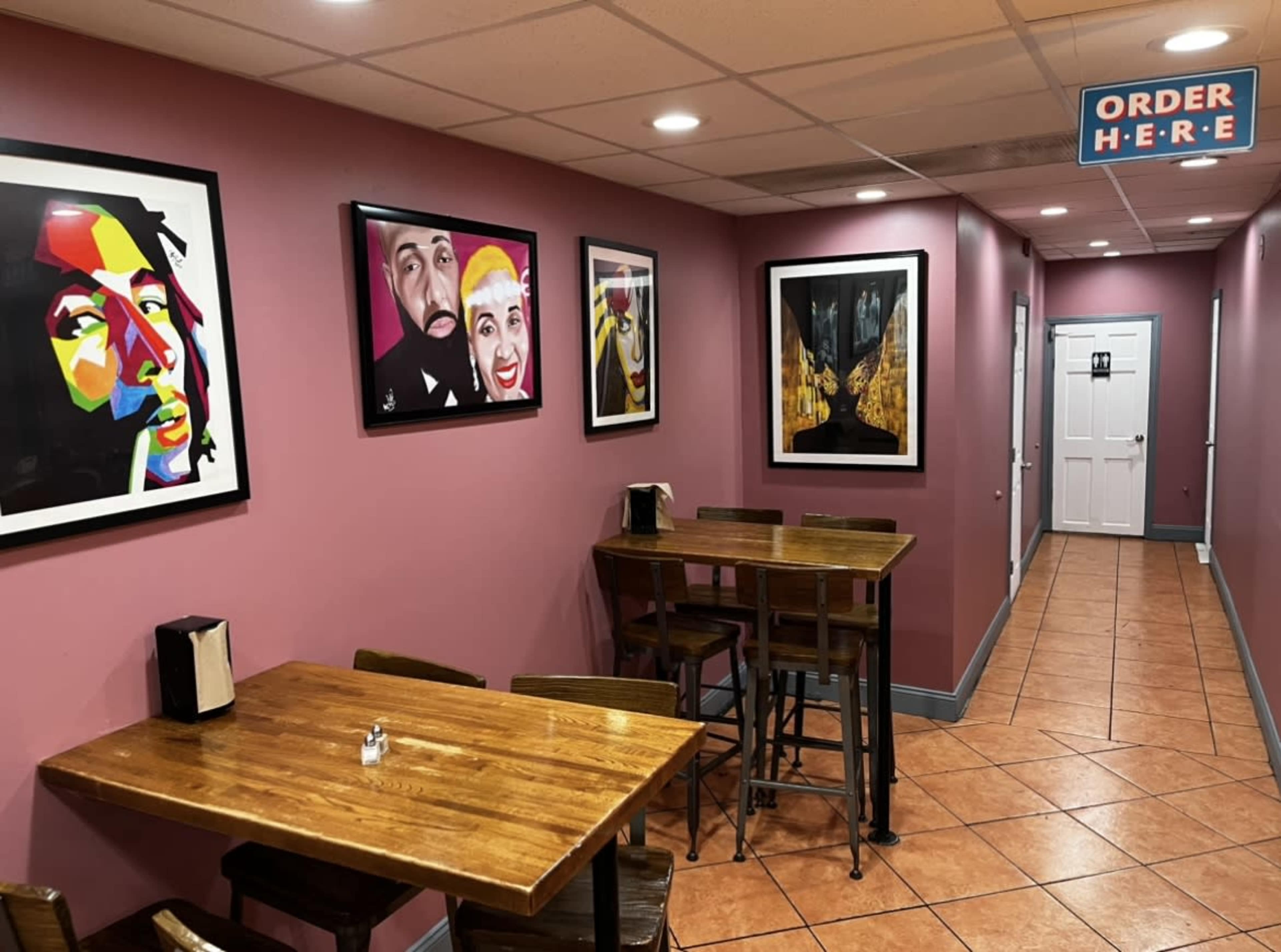 A corridor of a restaurant featuring two wooden tables, colorful artwork on the walls, and a sign indicating where to order.