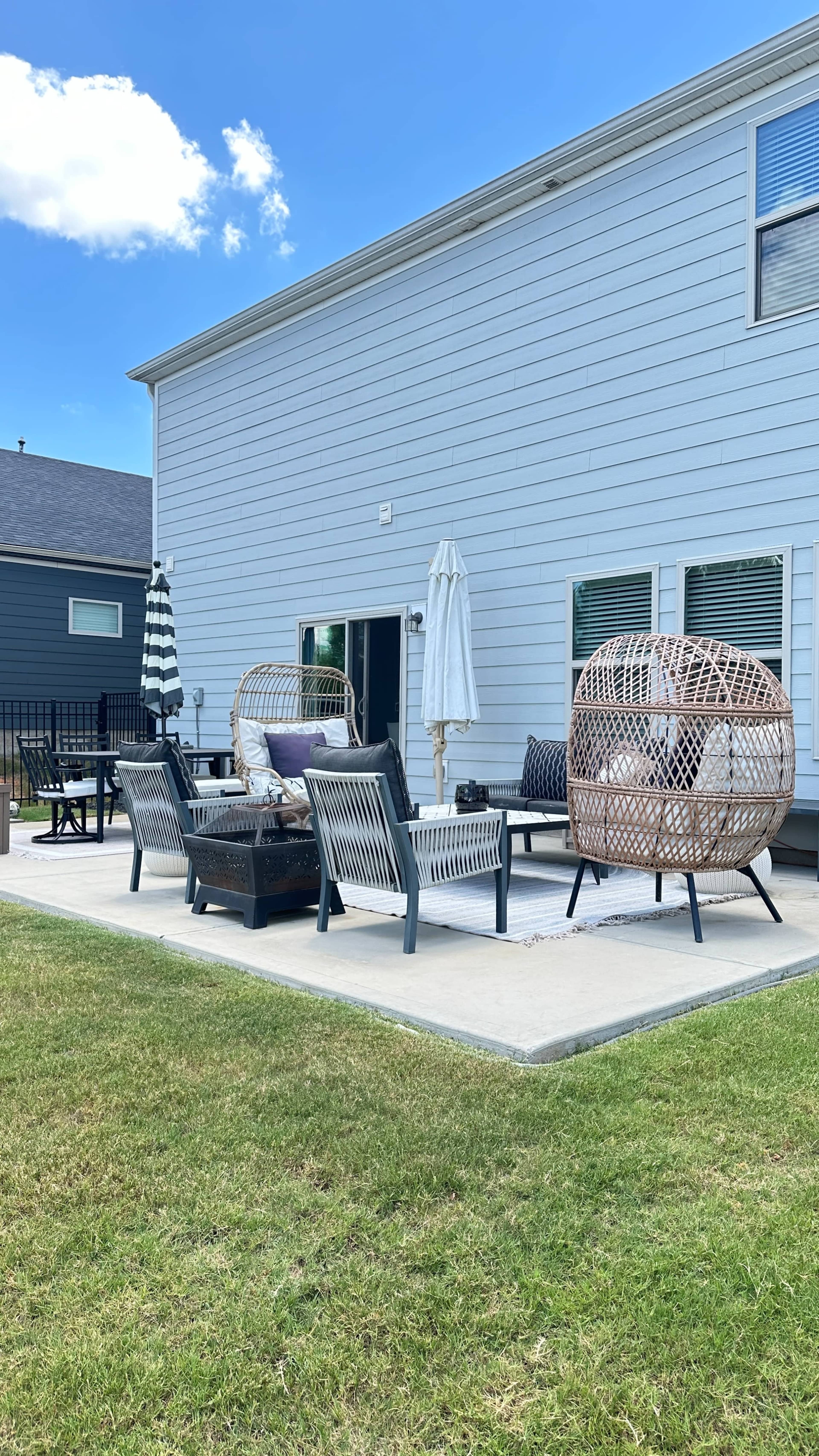 A patio area features various seating arrangements, including wicker chairs and a shaded umbrella, adjacent to a light blue house.
