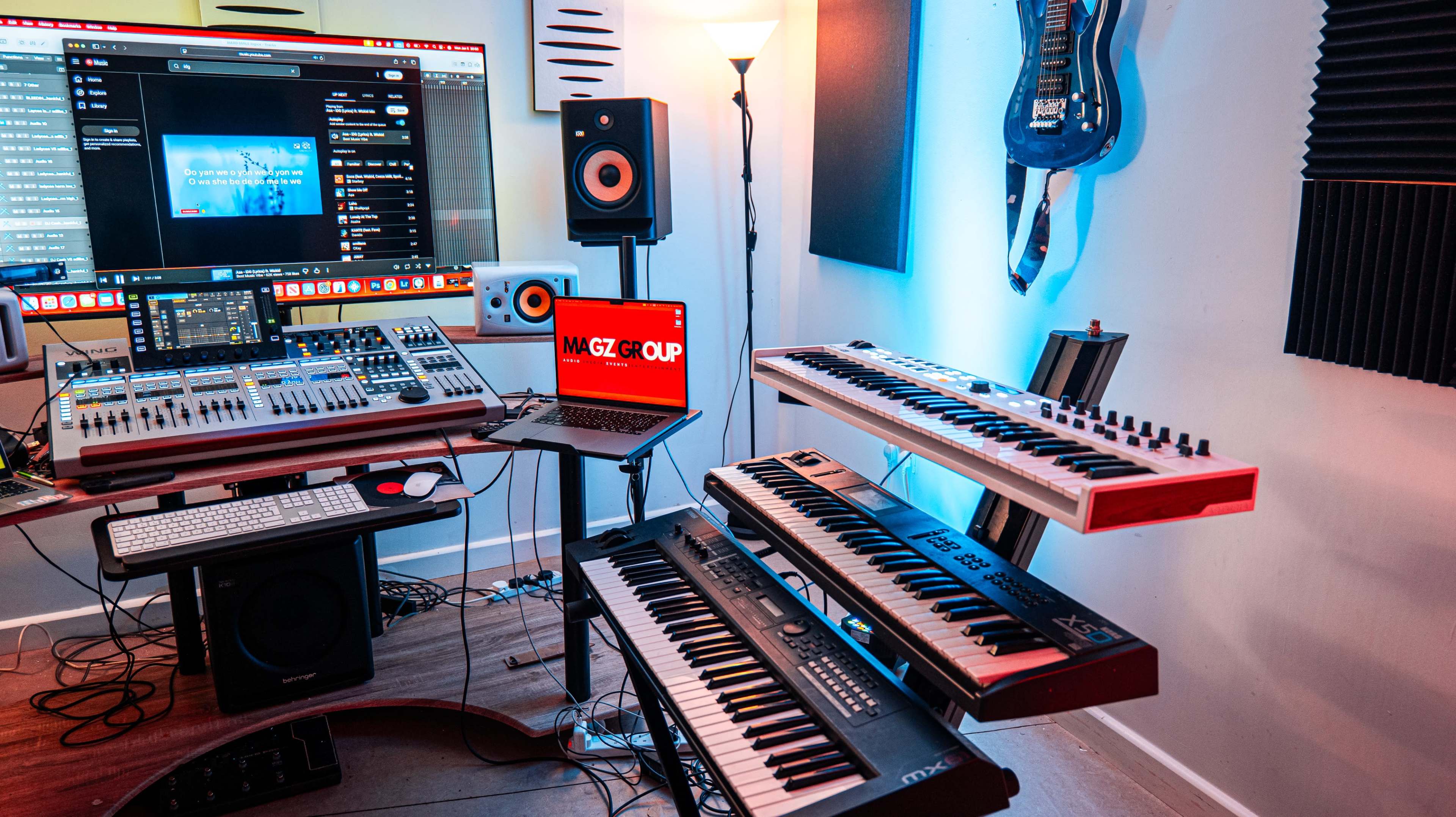 The image shows a home music studio featuring a variety of electronic equipment, including audio mixers, keyboards, a laptop, and a wall-mounted guitar.