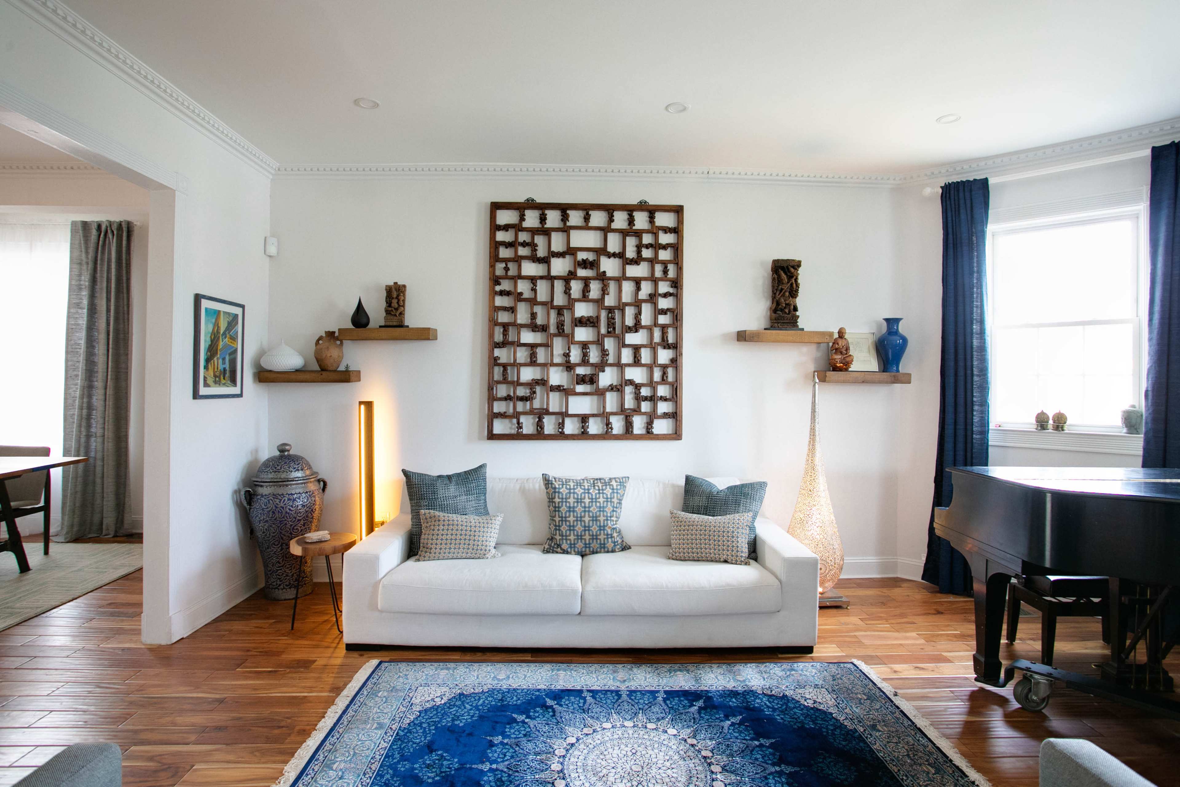 A white sofa with patterned pillows is positioned in a well-lit room featuring a wooden floor, decorative wall art, and plants, with a piano in the corner.
