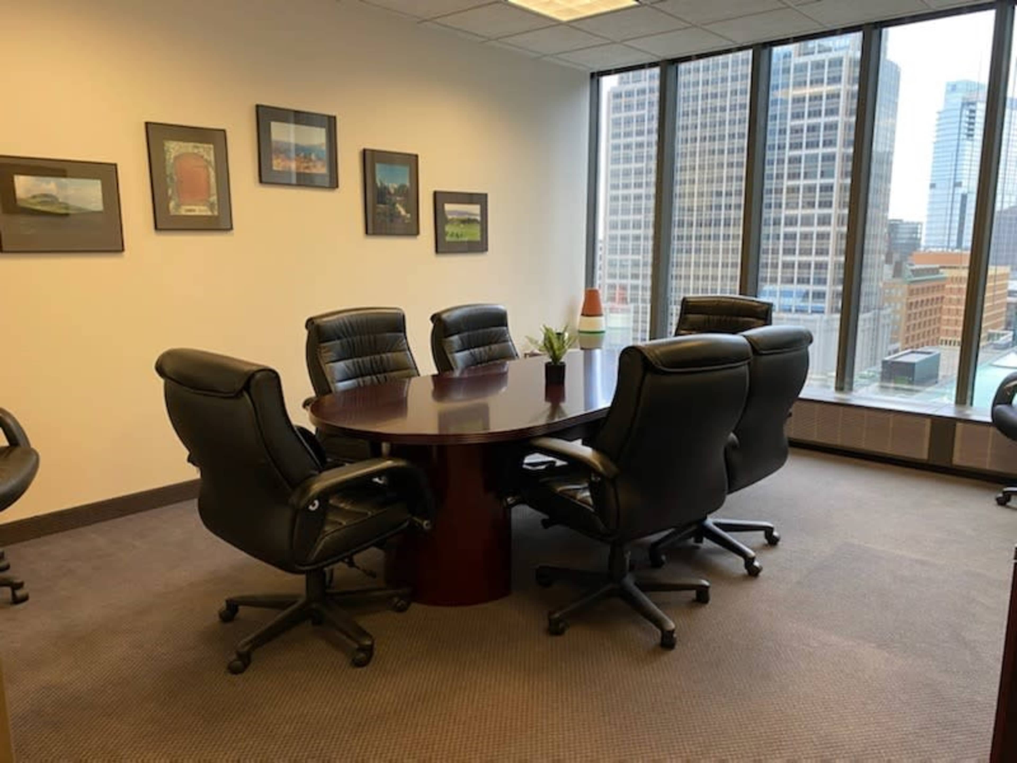 A round conference table surrounded by six black office chairs is positioned near large windows overlooking a cityscape.