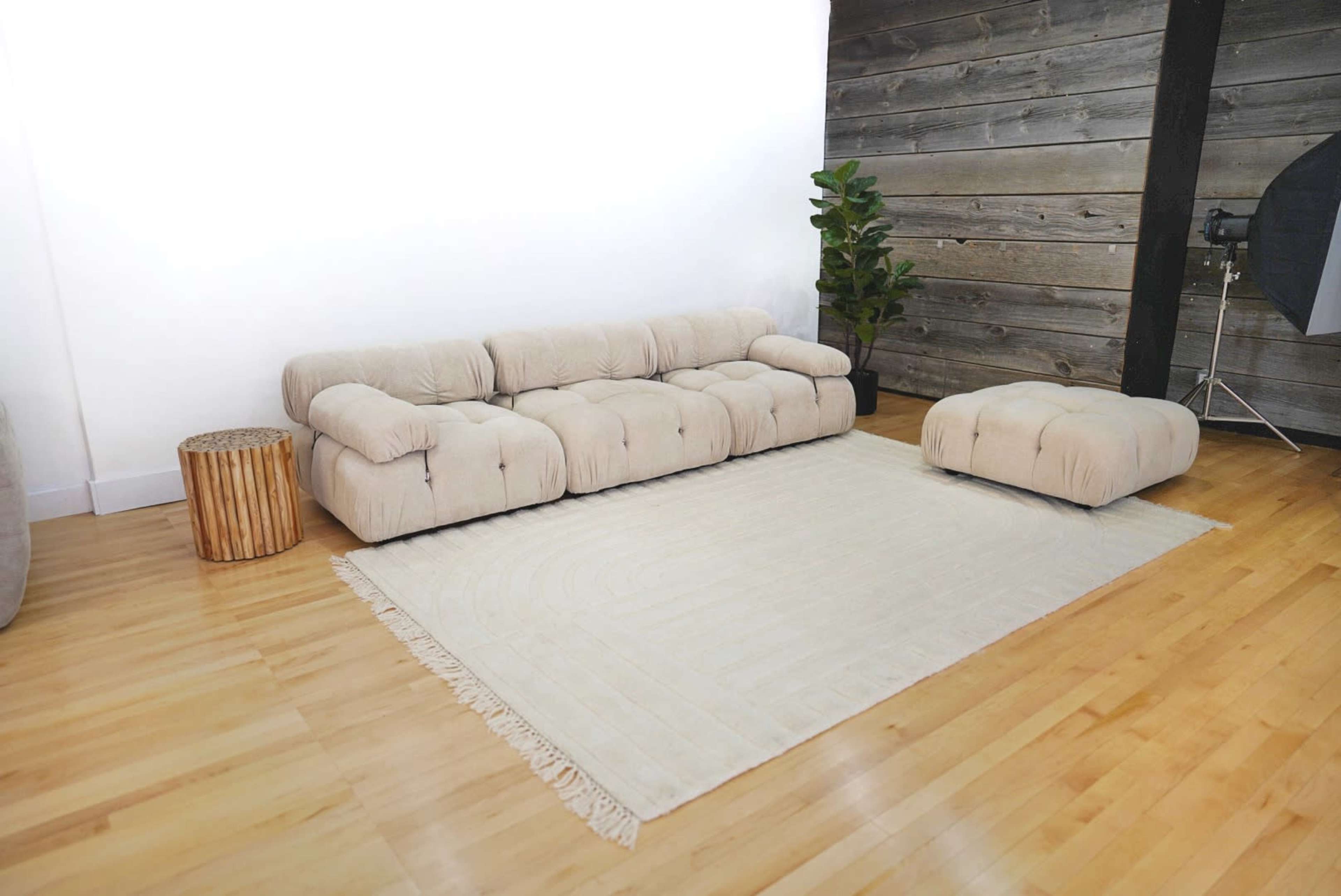 A light-colored sectional sofa and an ottoman on a large, beige rug in a spacious room with wooden flooring and a textured wall.