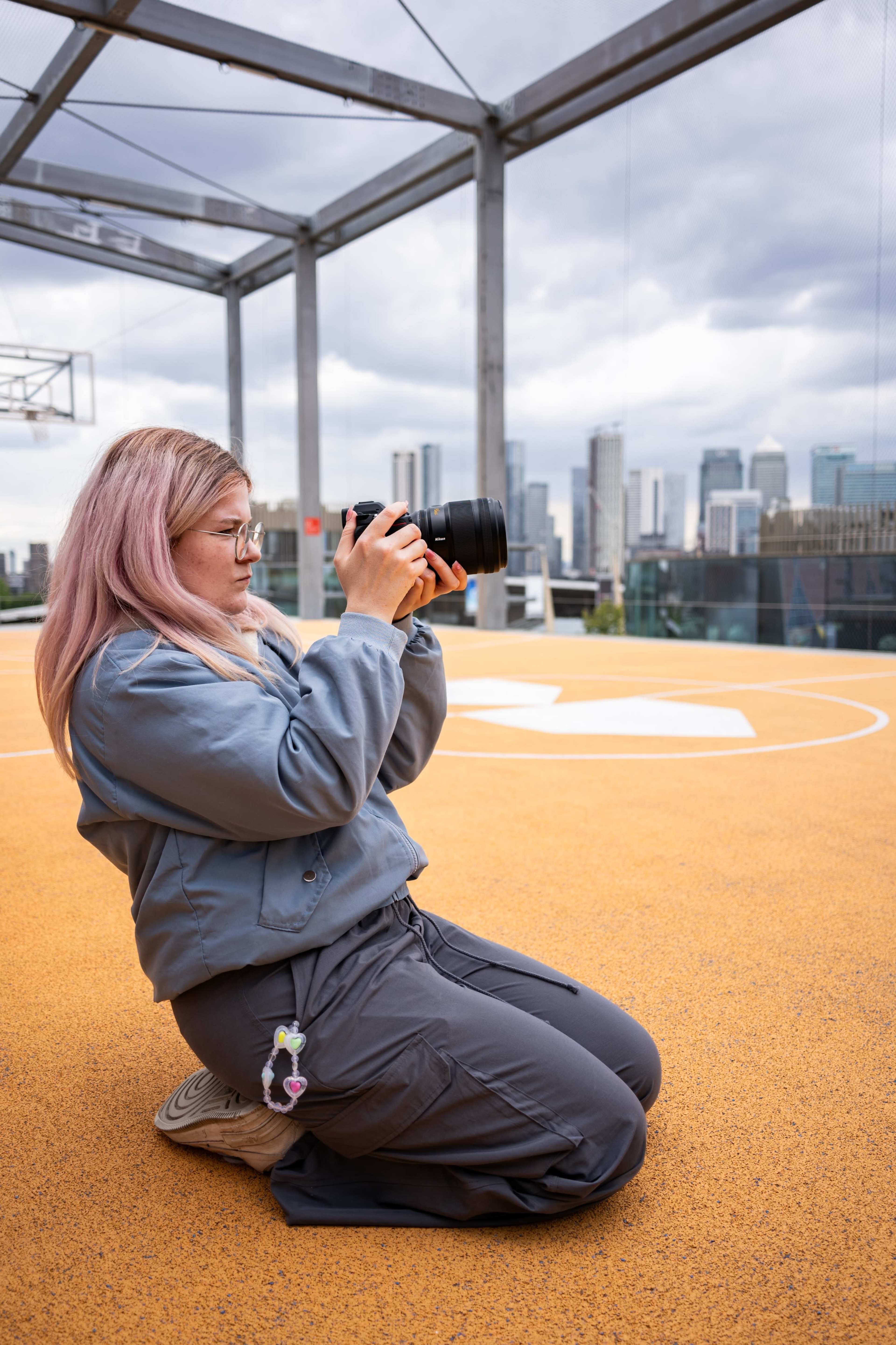 A woman with pink hair kneels on an orange basketball court while taking photos with a DSLR camera, featuring a city skyline in the background.