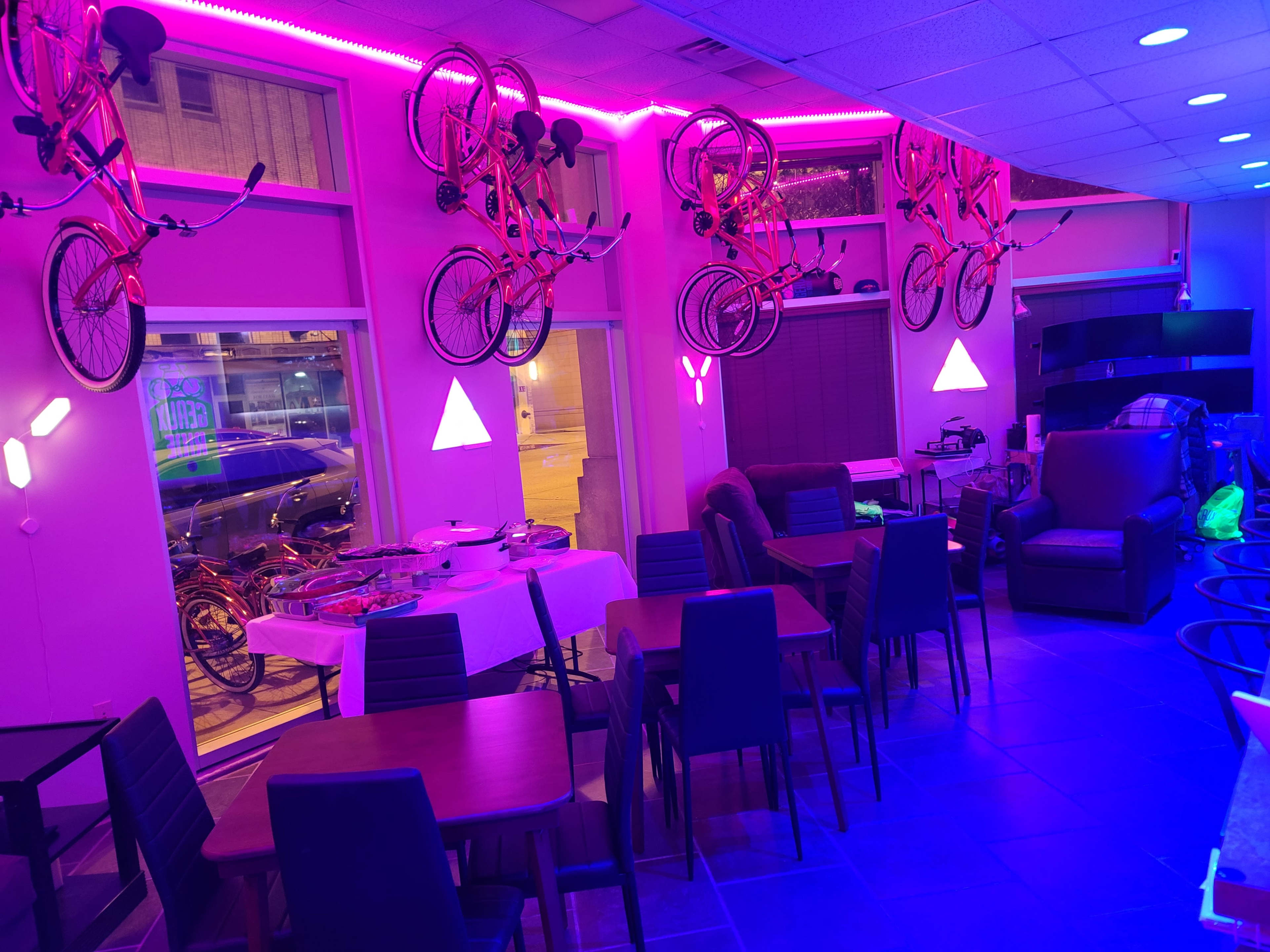 The interior of a cafe features tables and chairs arranged beneath bicycles mounted on the walls, illuminated by vibrant pink lighting.