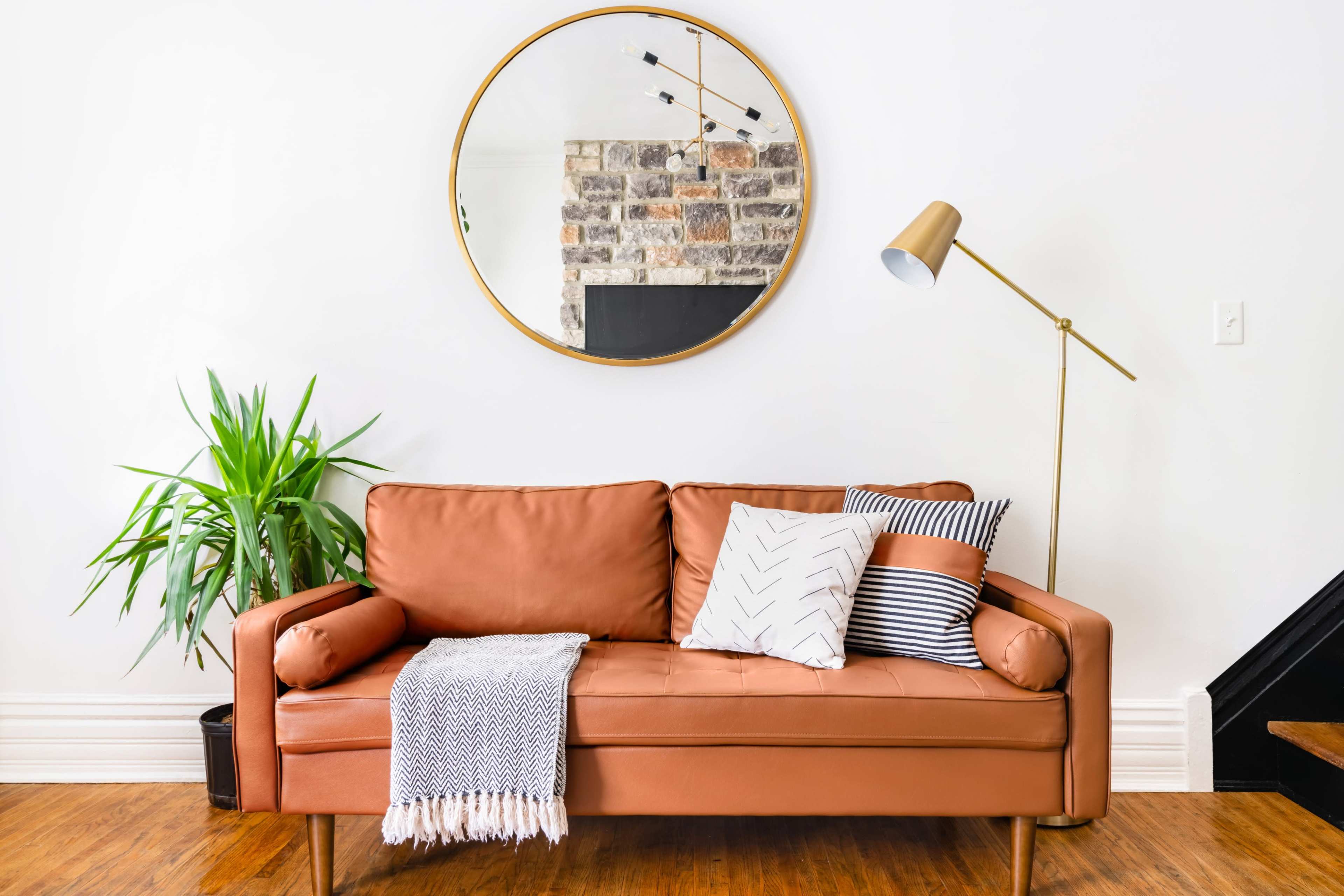 A brown leather sofa with patterned pillows is positioned in front of a circular mirror and a stone accent wall, accompanied by a small potted plant and a floor lamp.