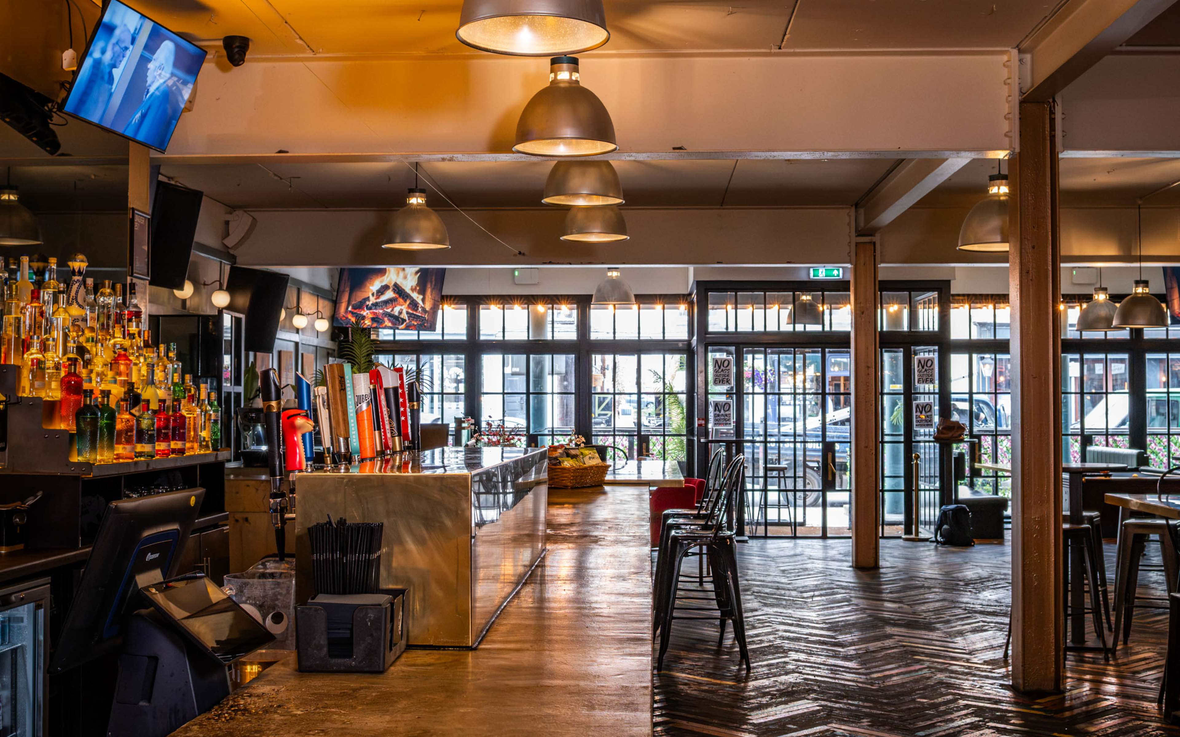 The image shows a spacious bar area with a long countertop, shelves filled with colorful liquor bottles, and large windows allowing natural light to enter.