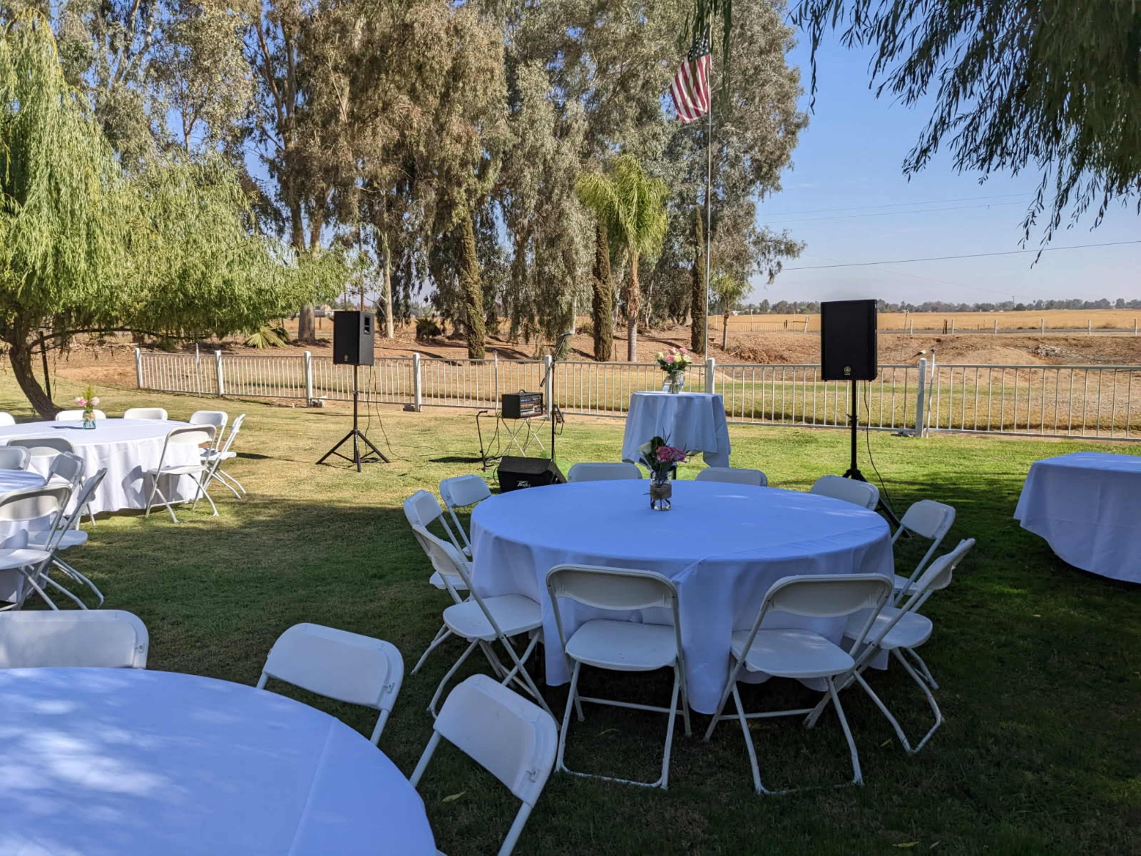 The scene shows a well-arranged outdoor venue with several round tables and white chairs set up on a green lawn, surrounded by trees and an open field in the background.