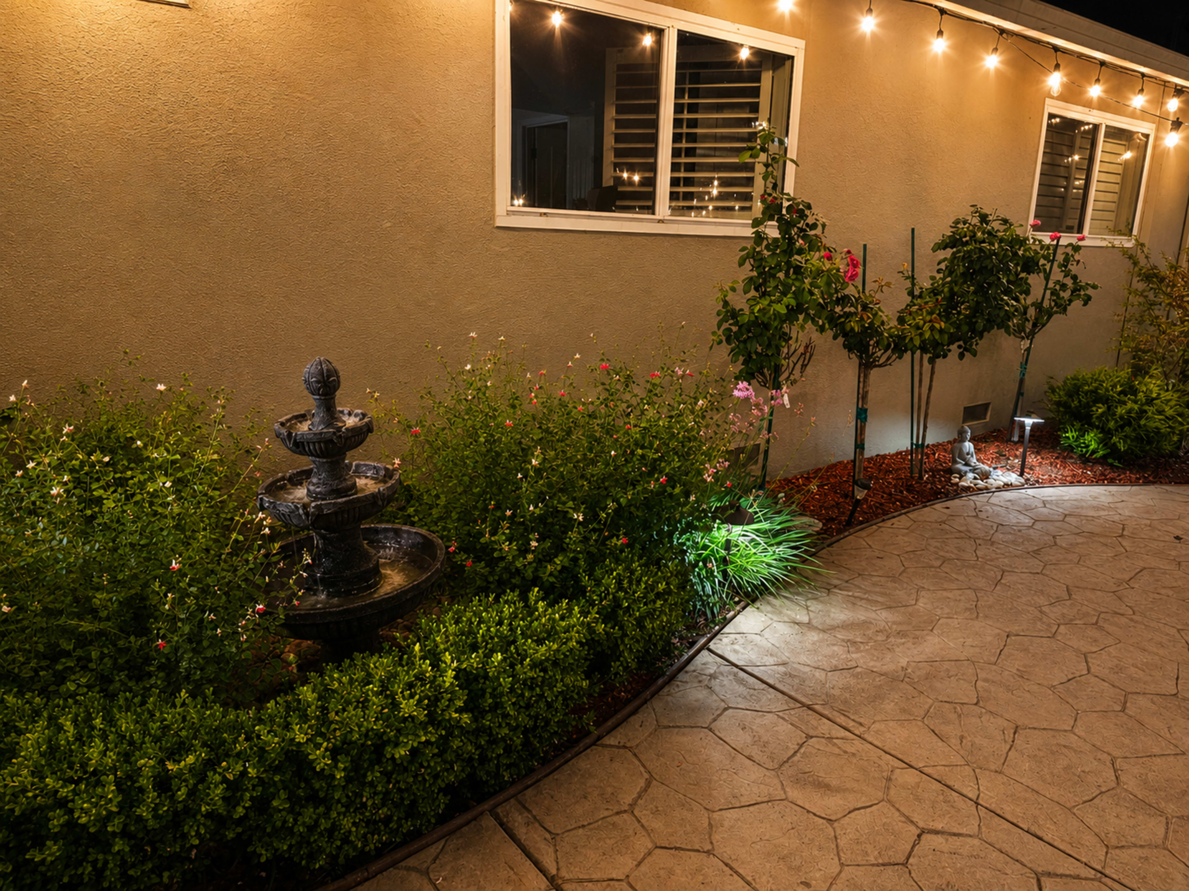 The image shows a garden path illuminated by string lights, featuring a decorative fountain surrounded by shrubs and flowers.