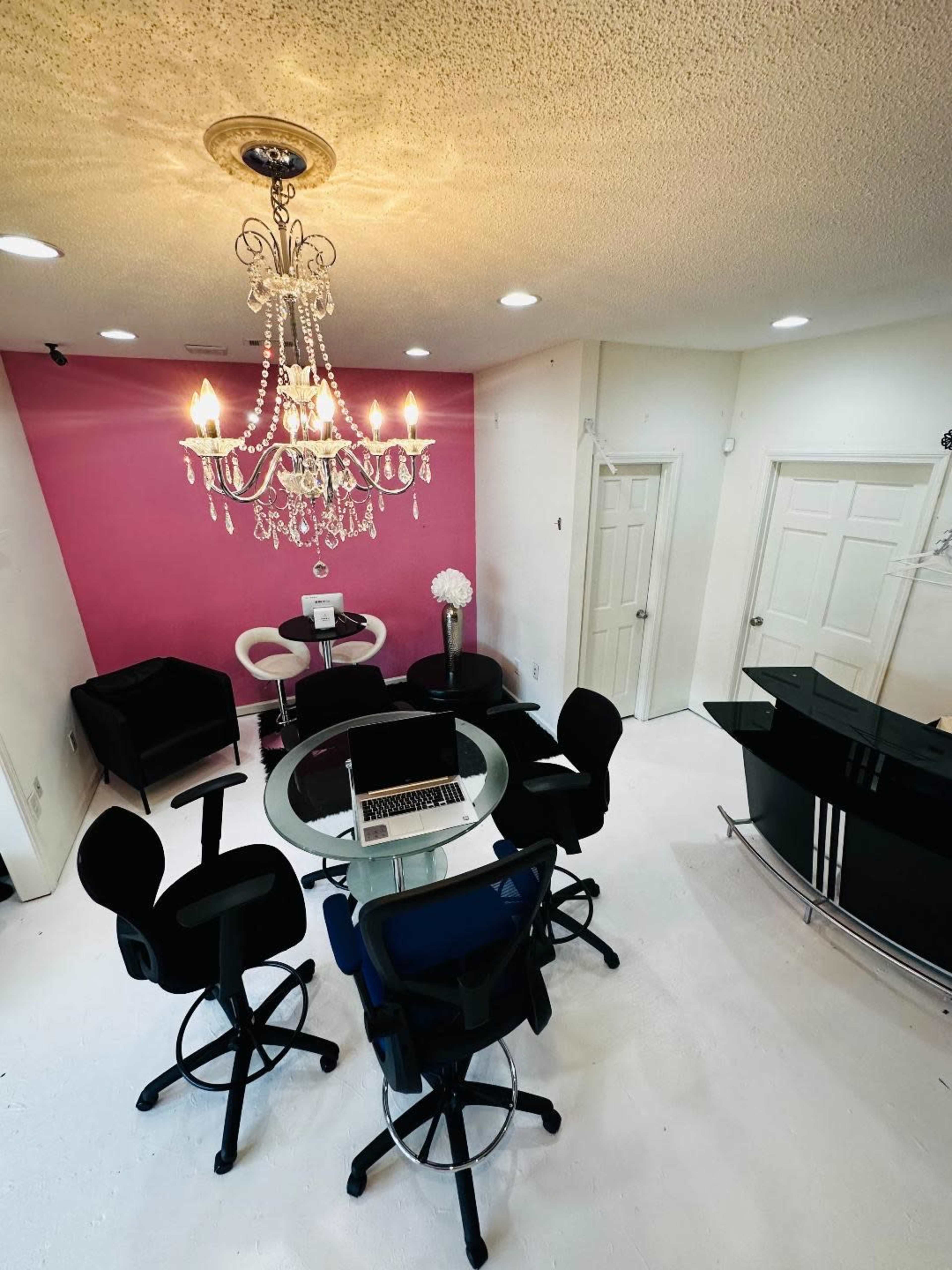 A well-lit salon features a glass table surrounded by high-backed chairs, with a chandelier above and pink walls accentuating the space.
