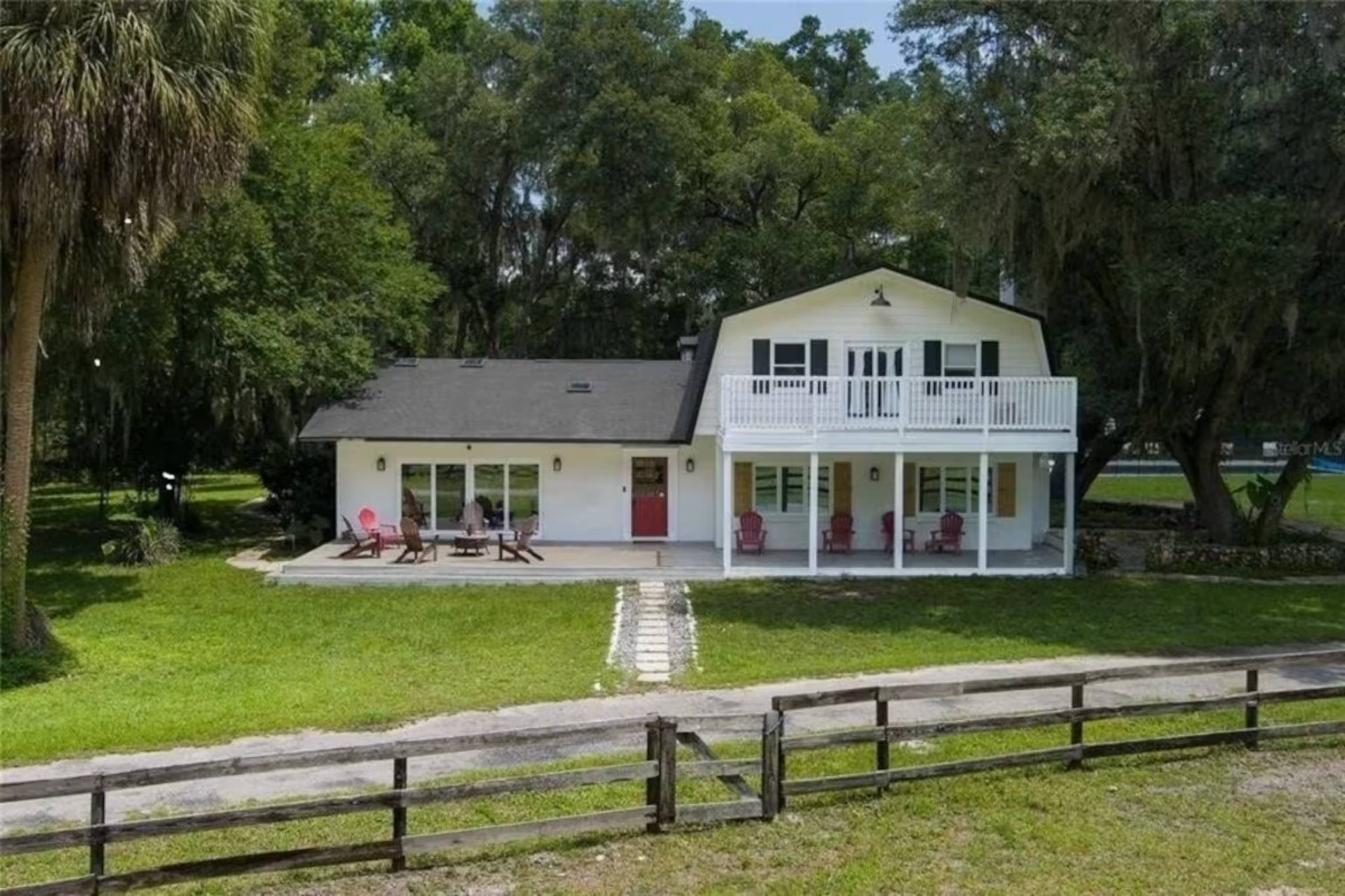 A two-story house with a large front porch and a balcony, surrounded by greenery and a wooden fence.