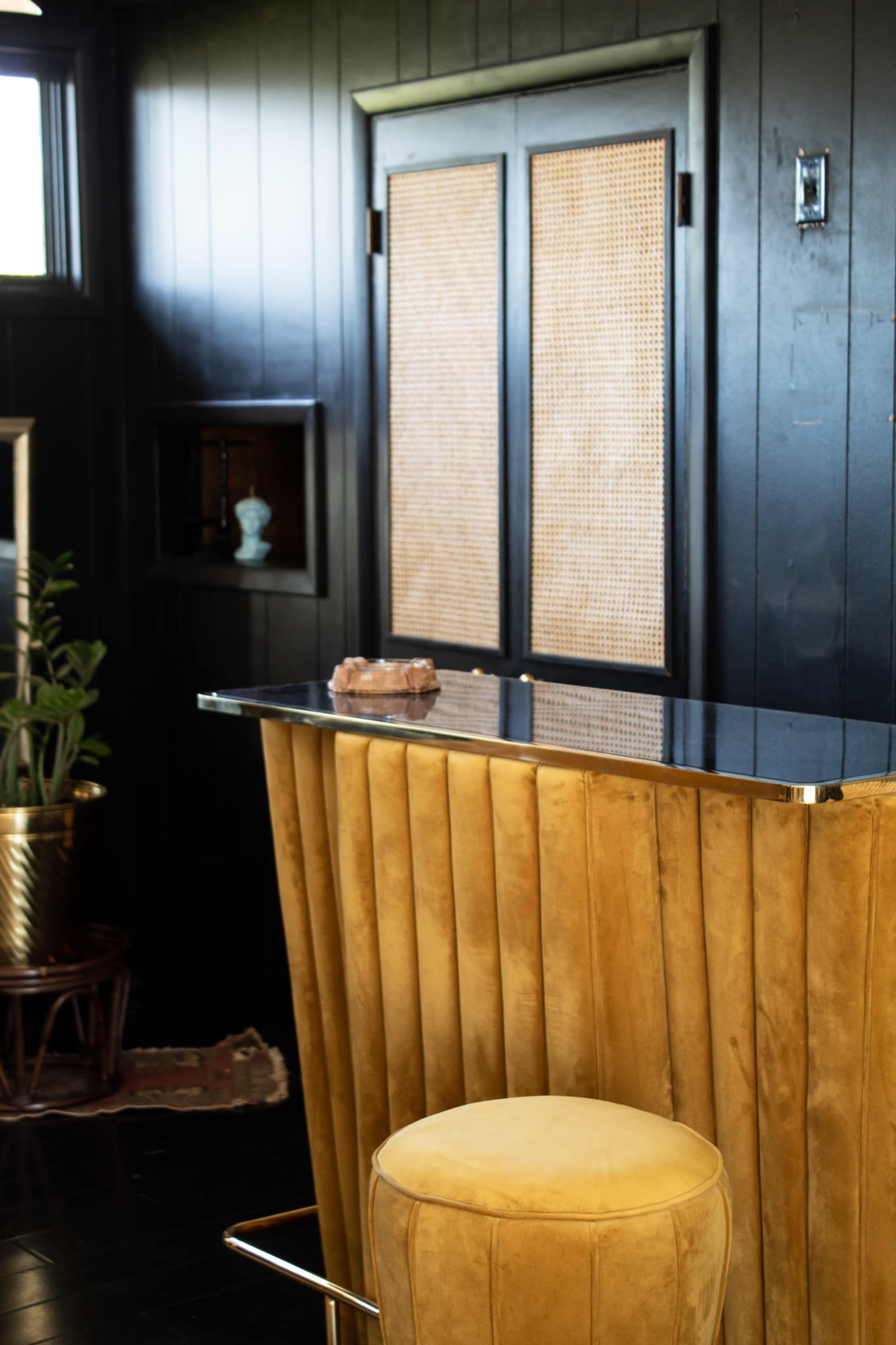 The image features a sleek bar area with a gold, pleated counter and a round stool, set against a black wall with wooden double doors.