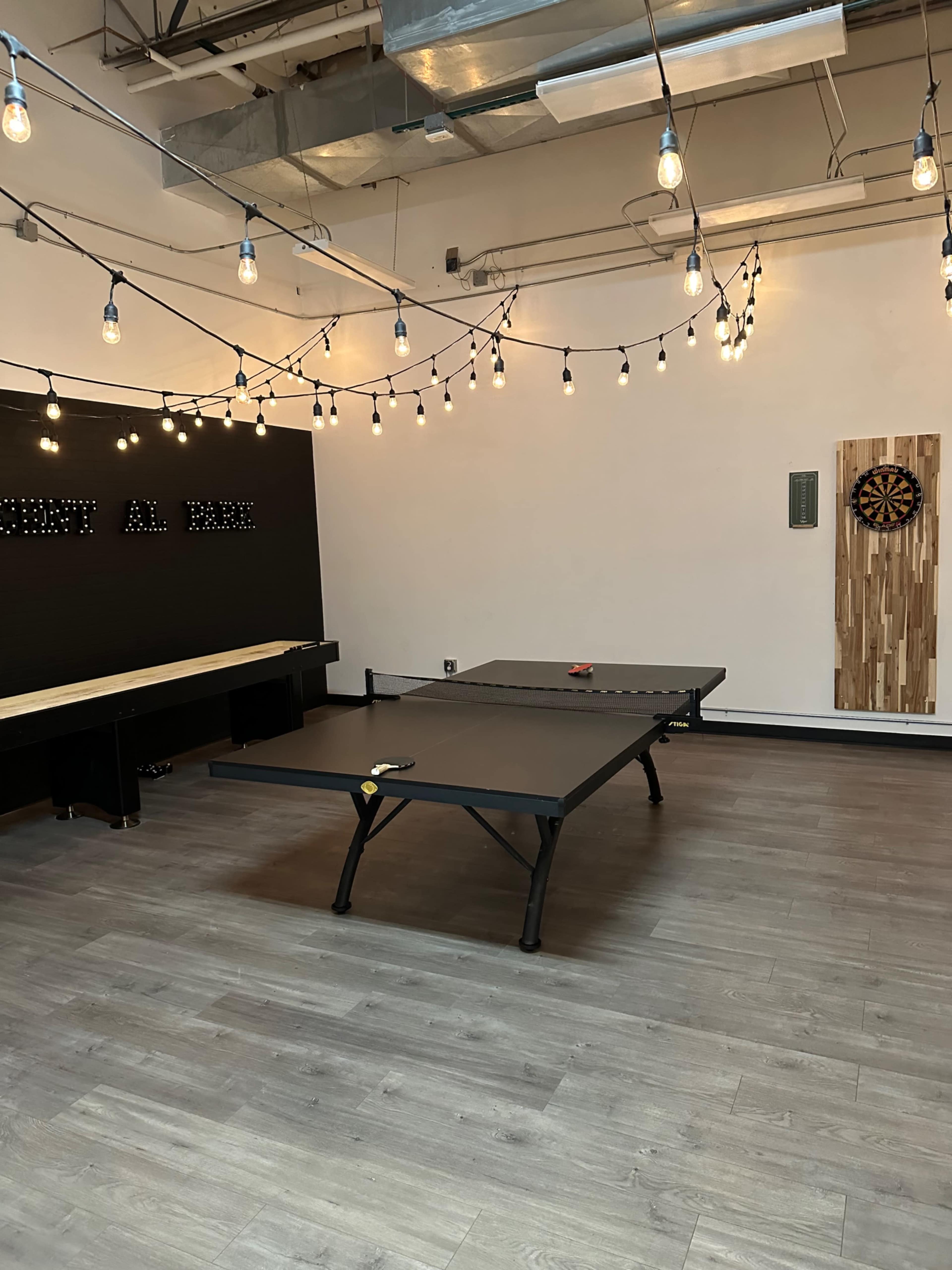 A recreational room with a ping pong table, a dartboard on a wooden panel, and string lights hanging from the ceiling.