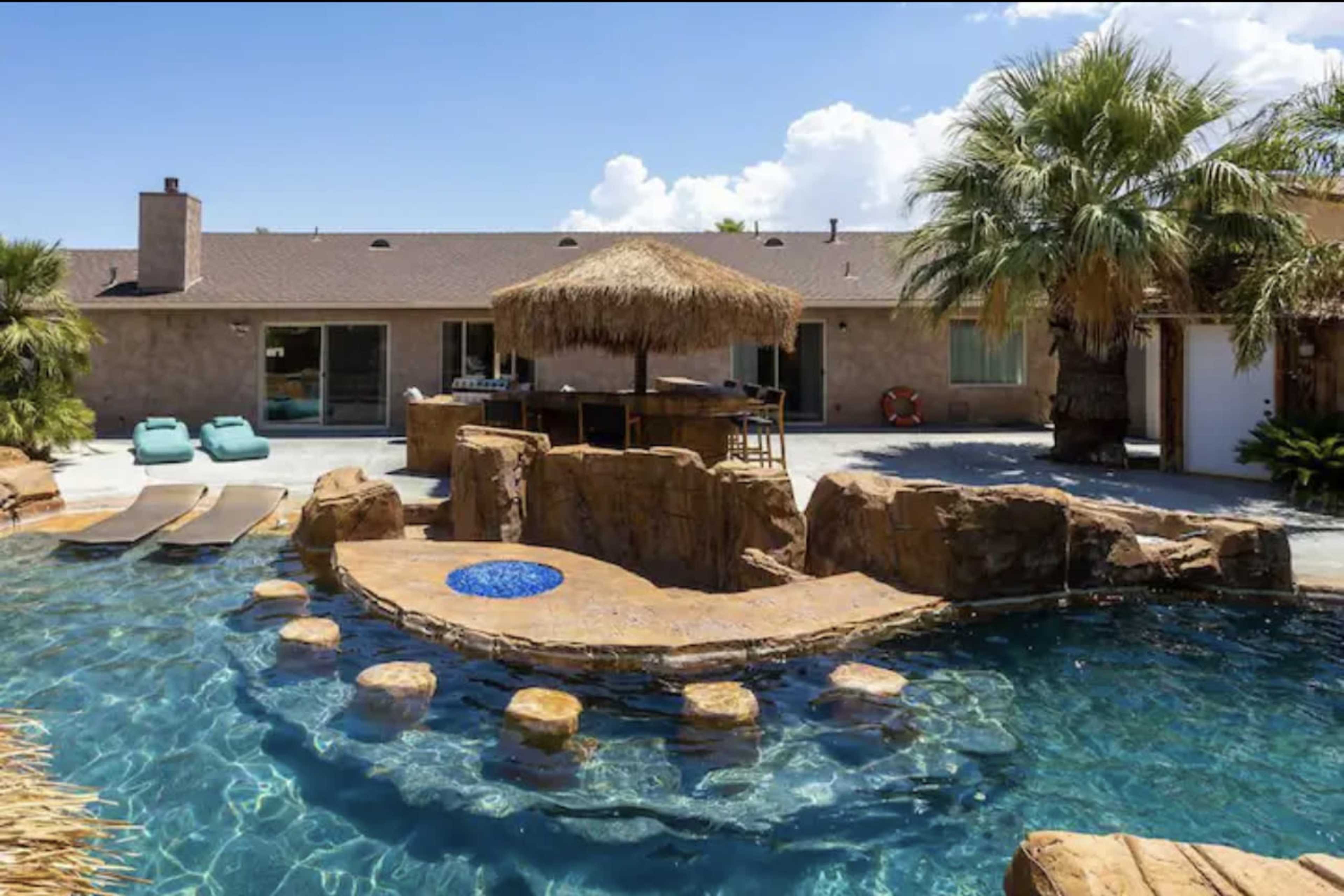 A backyard with a swimming pool featuring a stone formation and a tiki bar, surrounded by palm trees and lounge chairs.