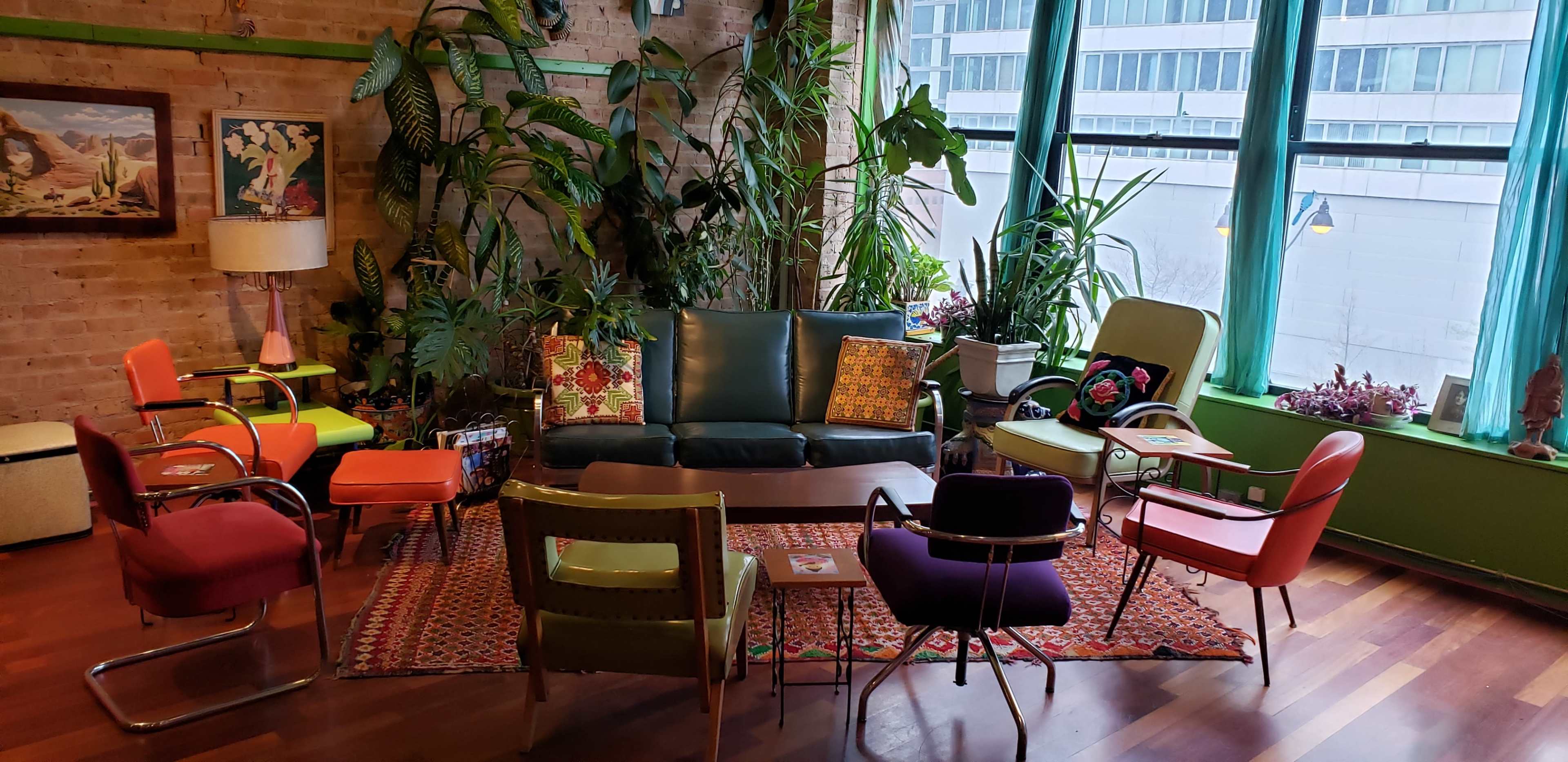A cozy sitting area features a green couch, colorful chairs, a patterned rug, and various plants against a backdrop of large windows.