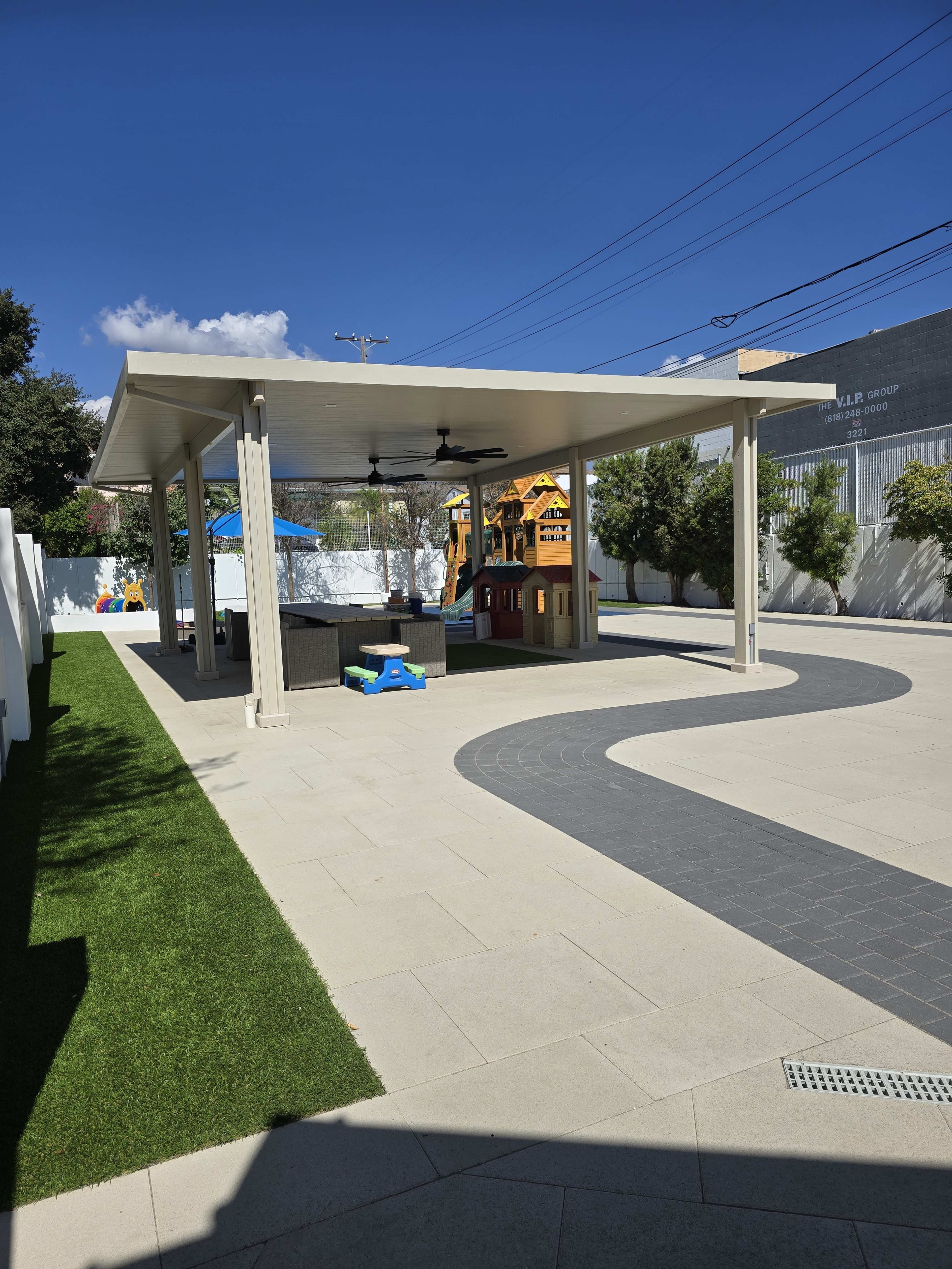 The image shows a spacious outdoor area featuring a covered pavilion with a playground in the background and a pathway leading through a grassy section.