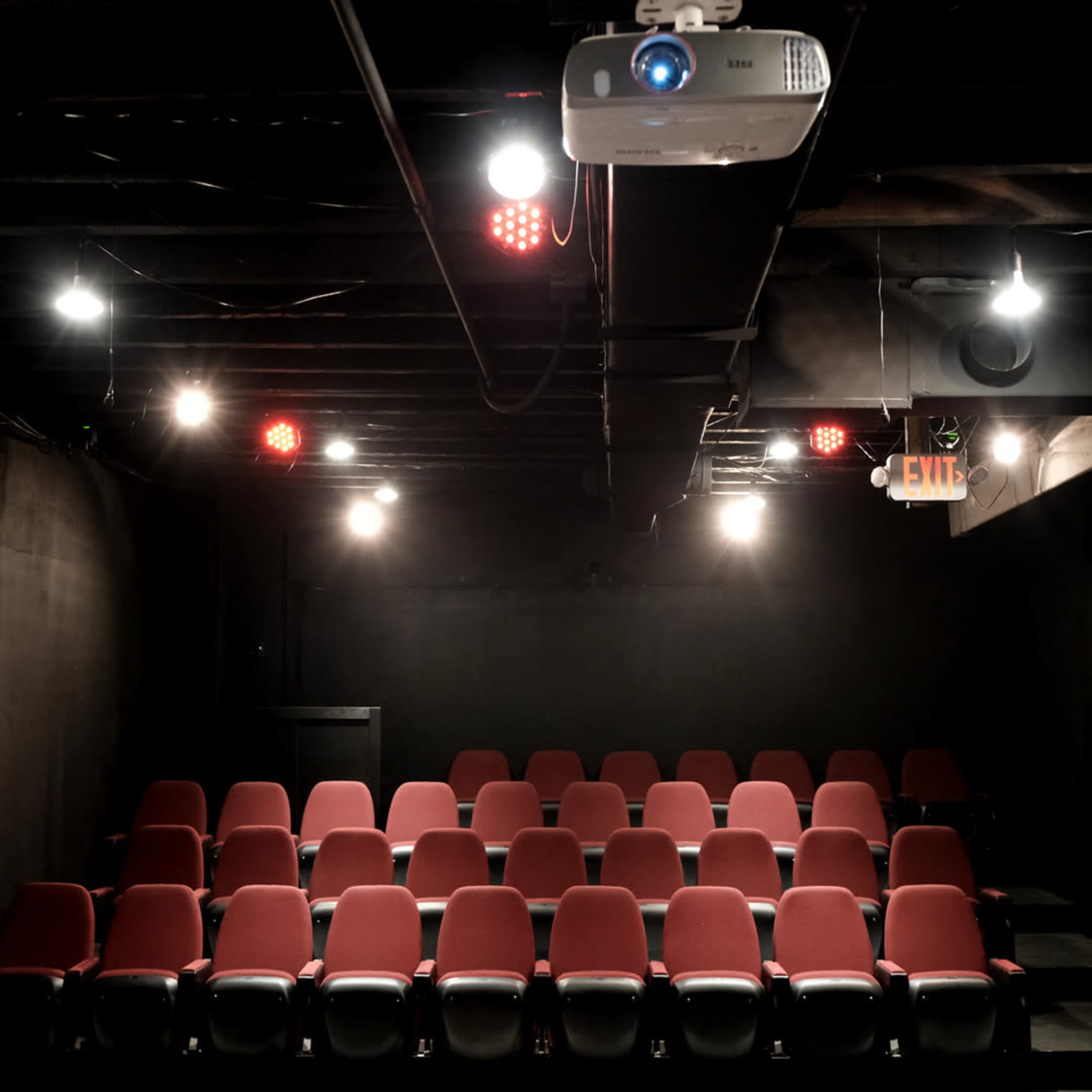 A small theater with red seats arranged in rows and a ceiling-mounted projector above.
