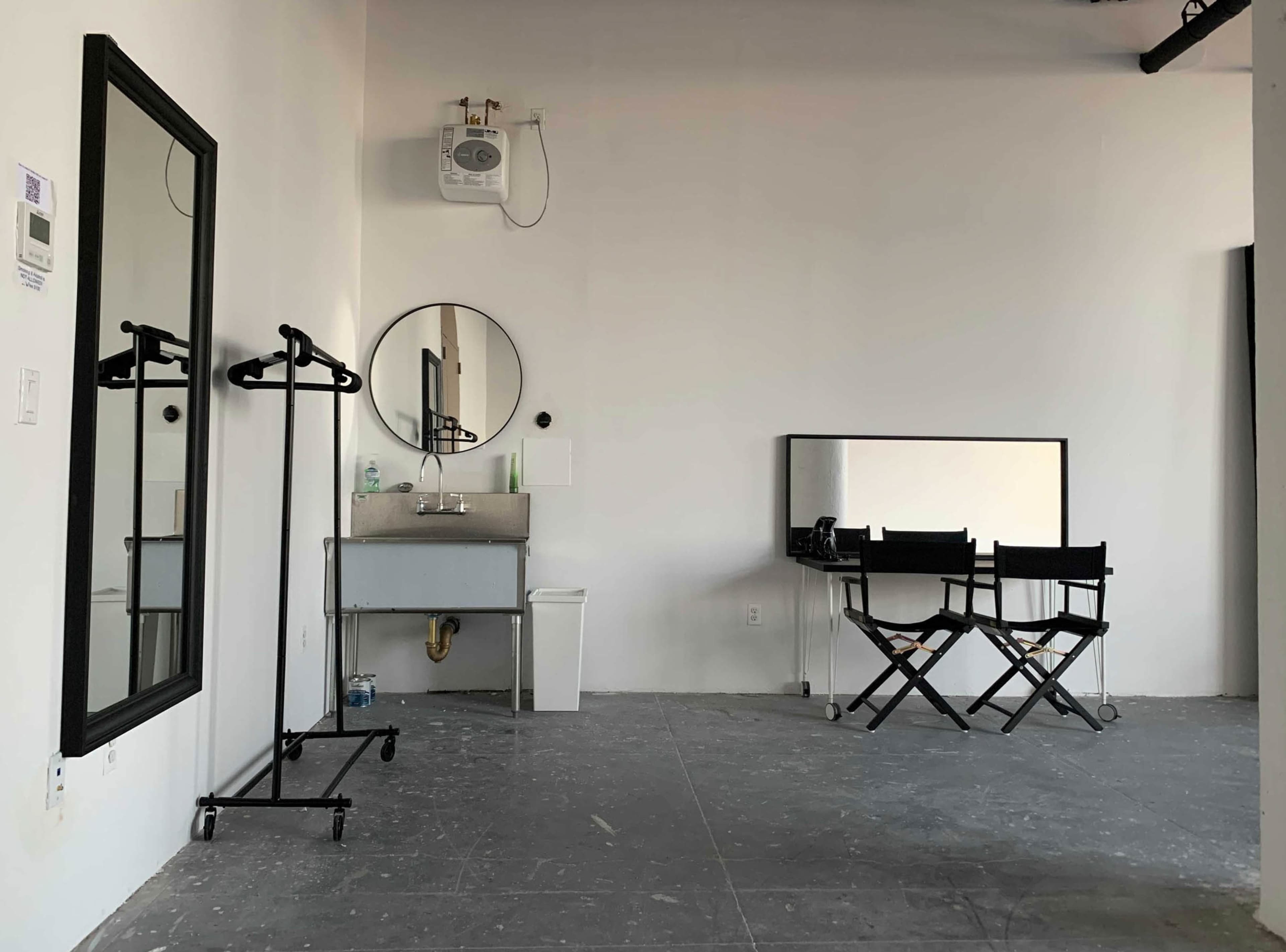 The scene shows a minimalist room with a large mirror, a small sink, two director's chairs, and a clothing rack against a white wall.