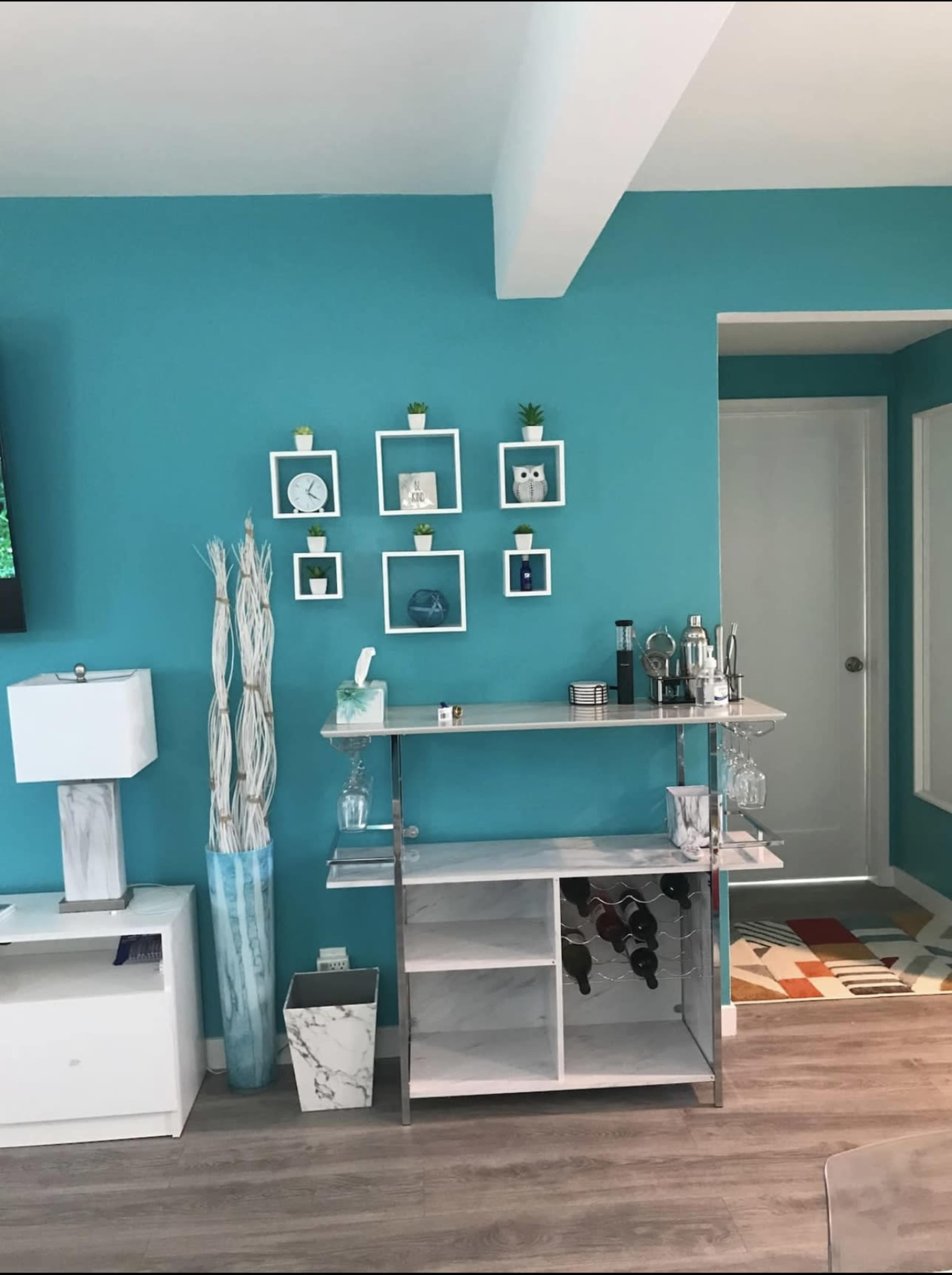 The image shows a turquoise wall featuring a shelf with decorative items, a bar cart with wine storage, and a lamp beside it.