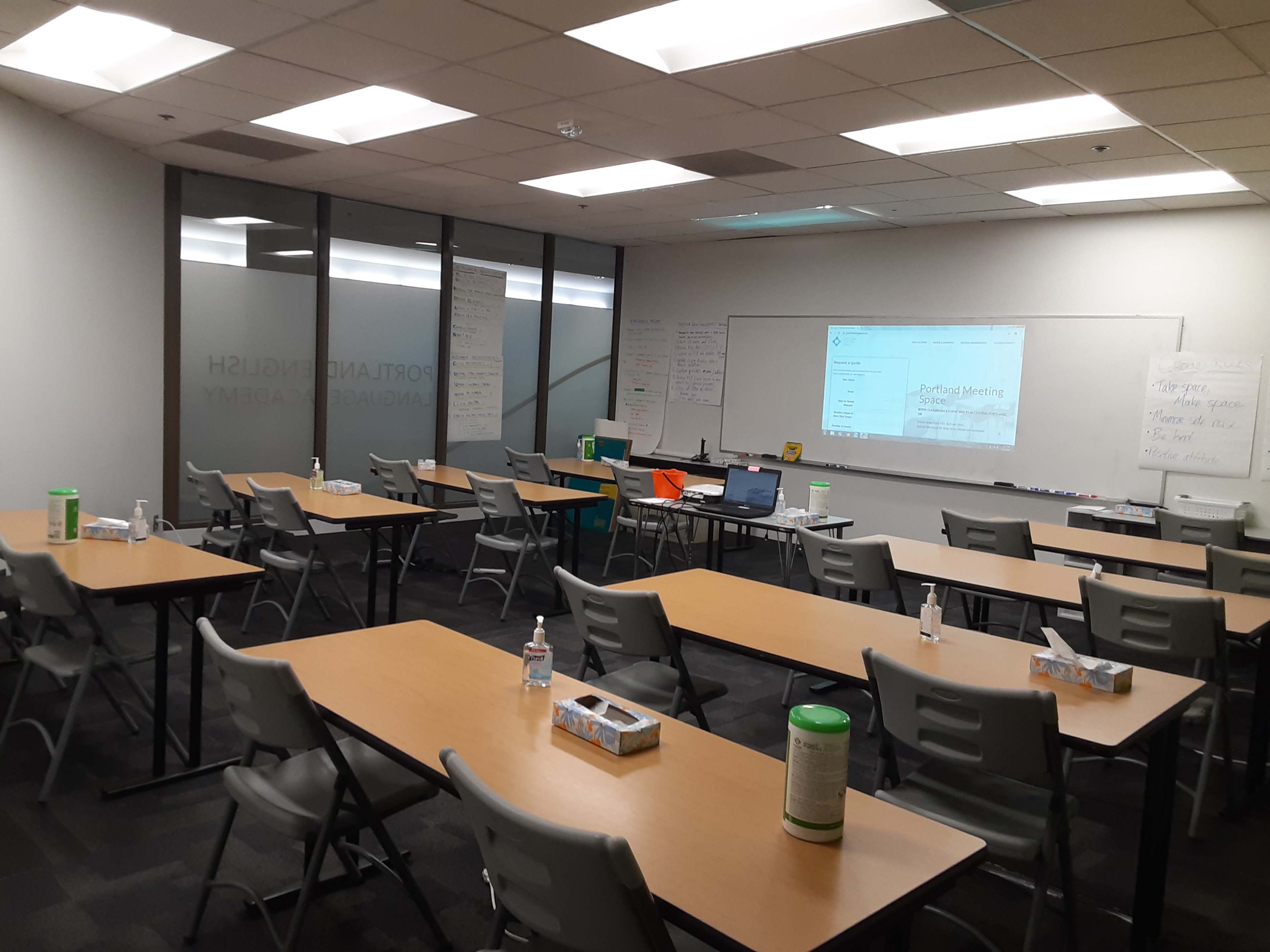 A classroom or training room with several tables and chairs arranged in rows, a projector displaying content on a screen, and whiteboards on the walls.