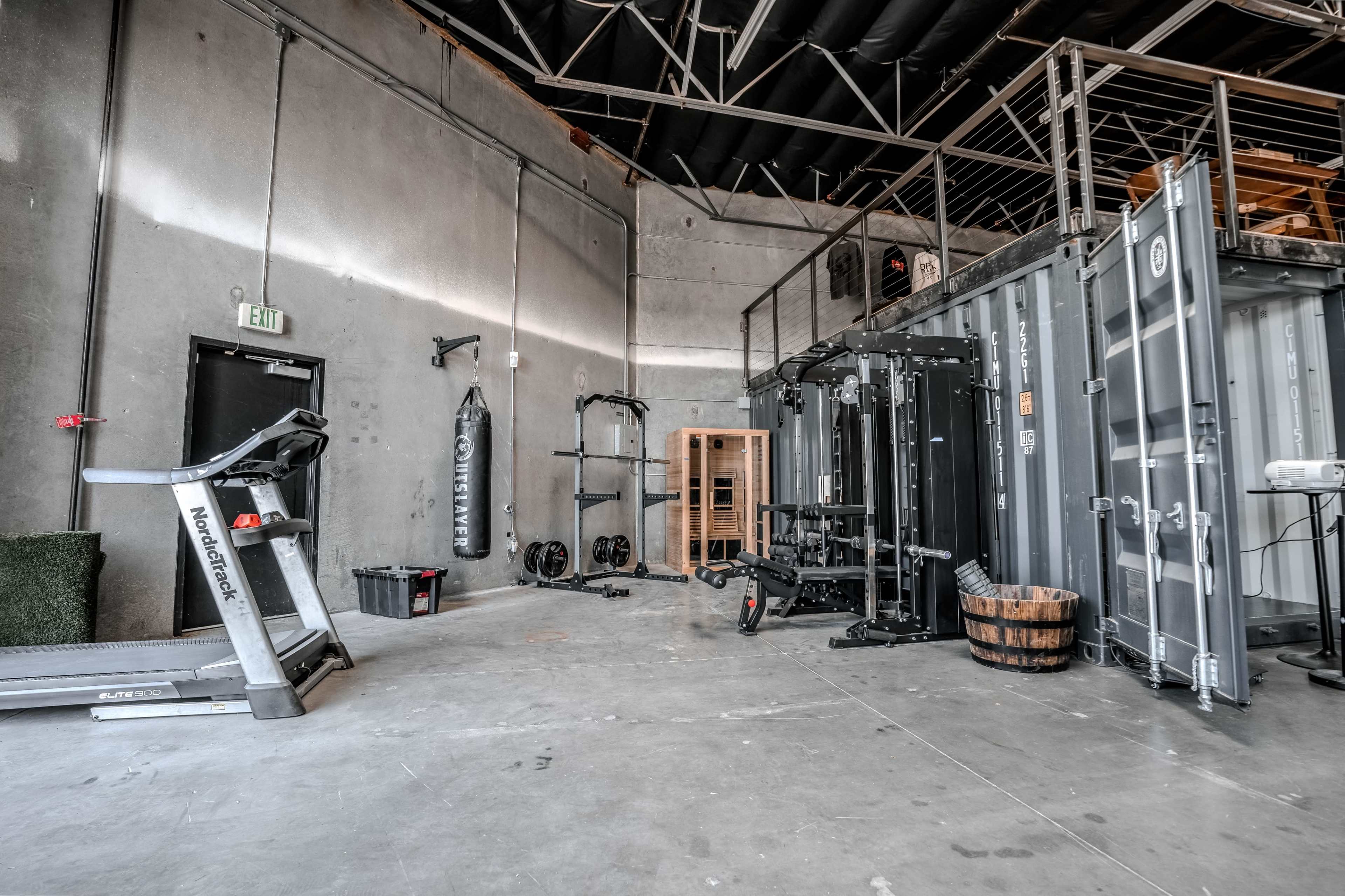 The image shows a gym space with various exercise equipment, including a treadmill, a weightlifting station, and a punching bag, set against concrete walls and a mezzanine level.