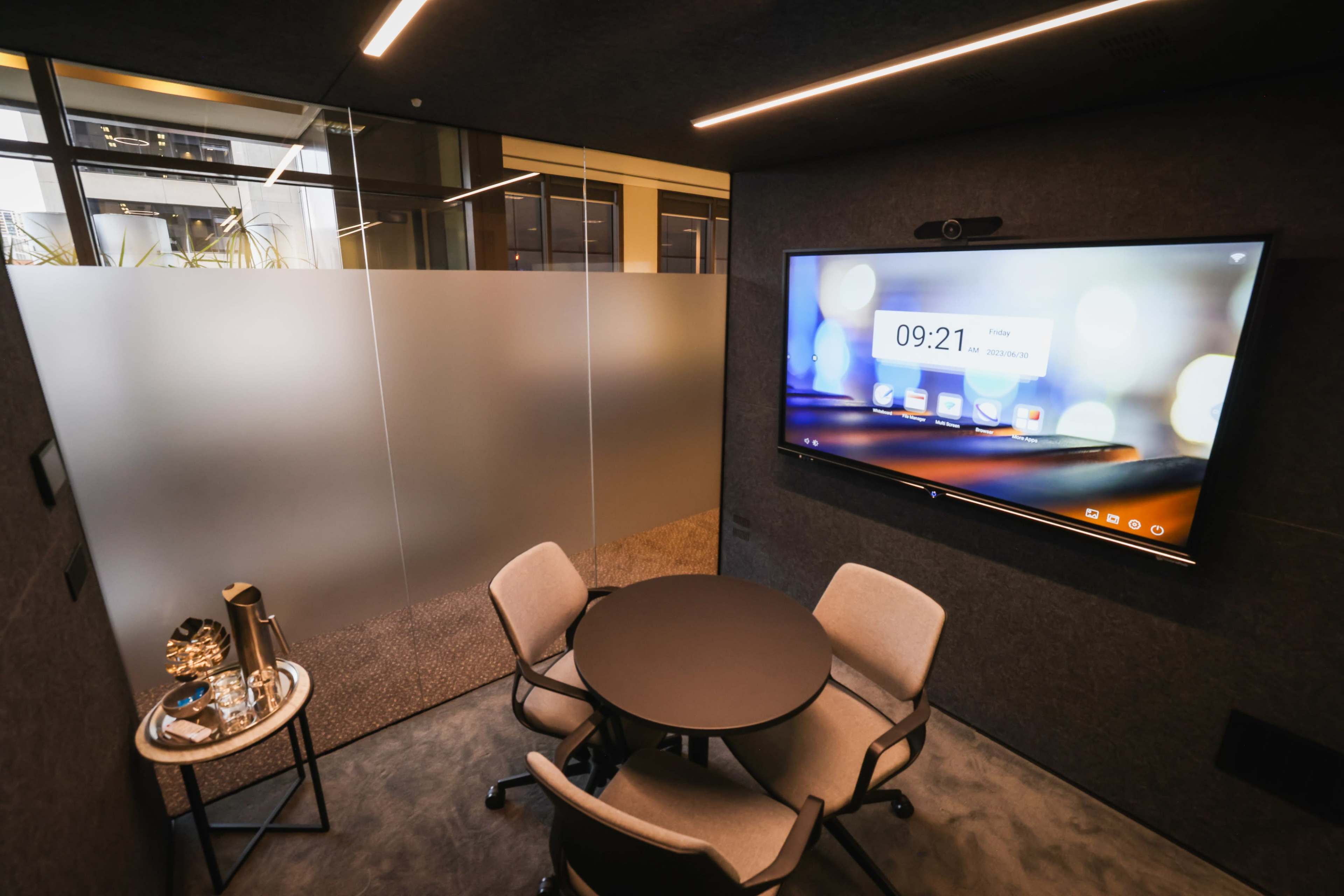The image shows a small, modern conference room with a round table, two chairs, and a wall-mounted screen displaying the time and app icons.