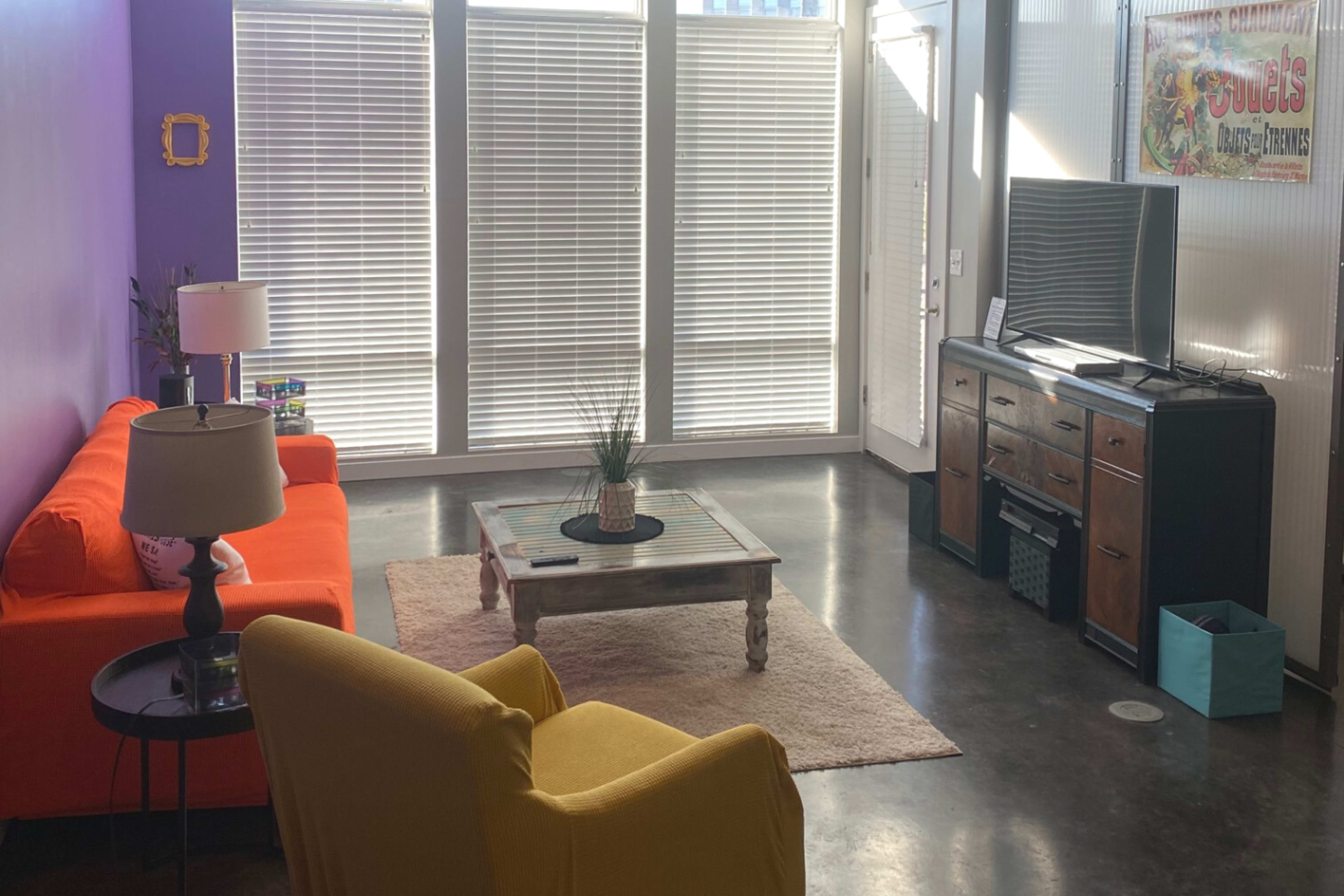 A living room with an orange sofa, a yellow armchair, a wooden coffee table, and a television on a dark wooden cabinet, all in front of large windows with blinds.