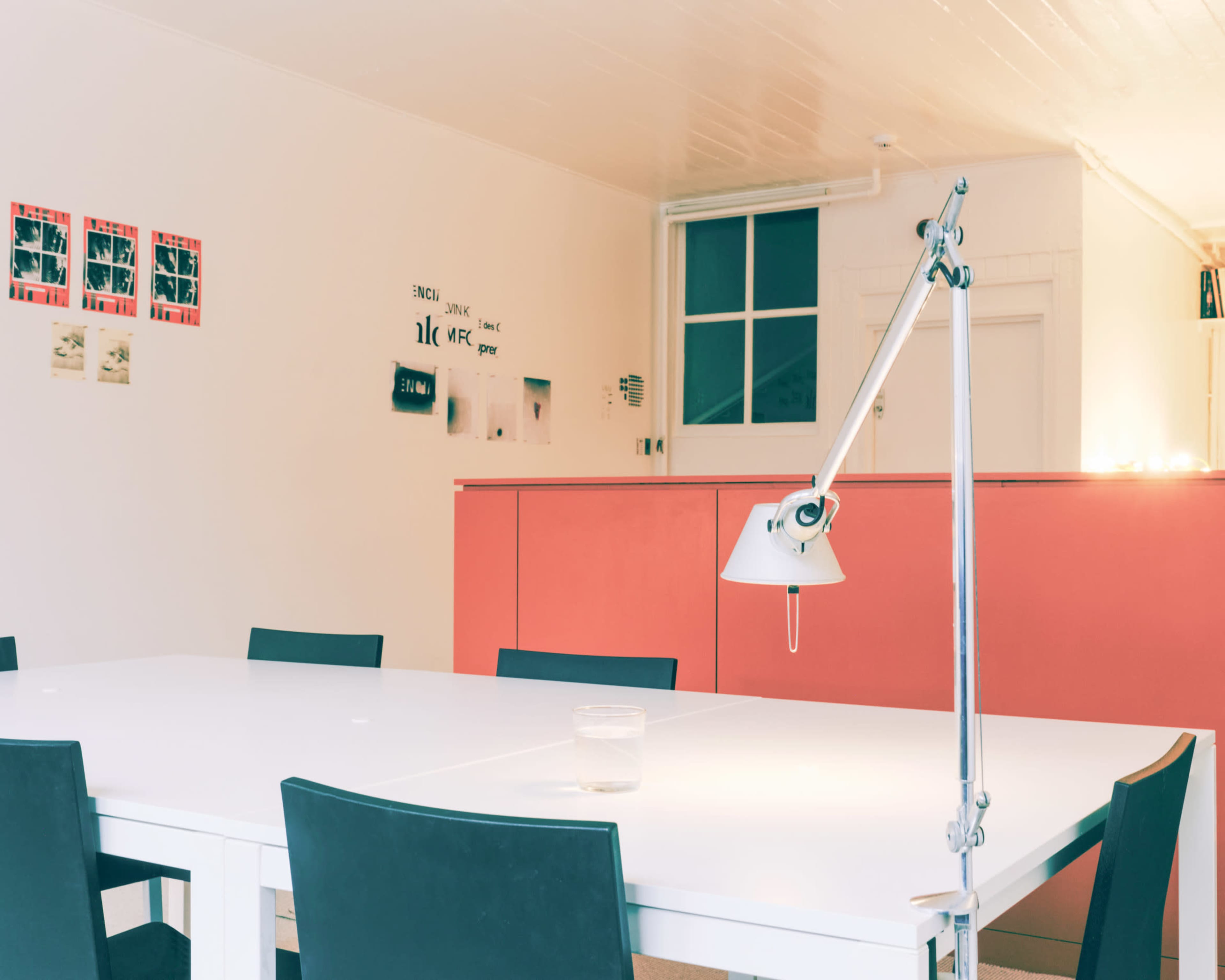 A bright, modern meeting room features a large white table surrounded by black chairs, with a lamp and a coral-colored wall in the background adorned with framed photographs.