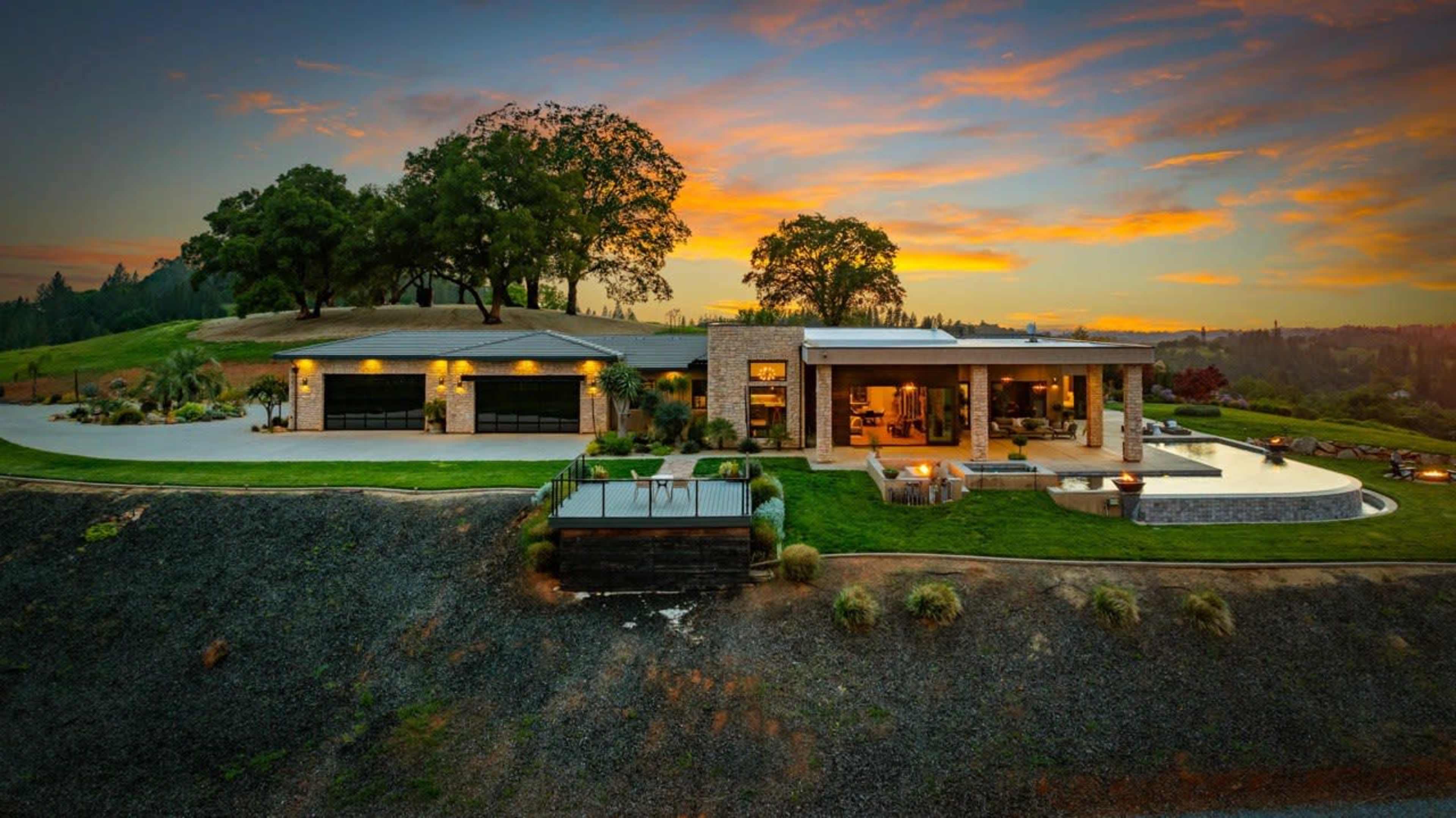 A modern house with large windows is situated on a hillside, surrounded by trees and grass, against a backdrop of a colorful sunset.