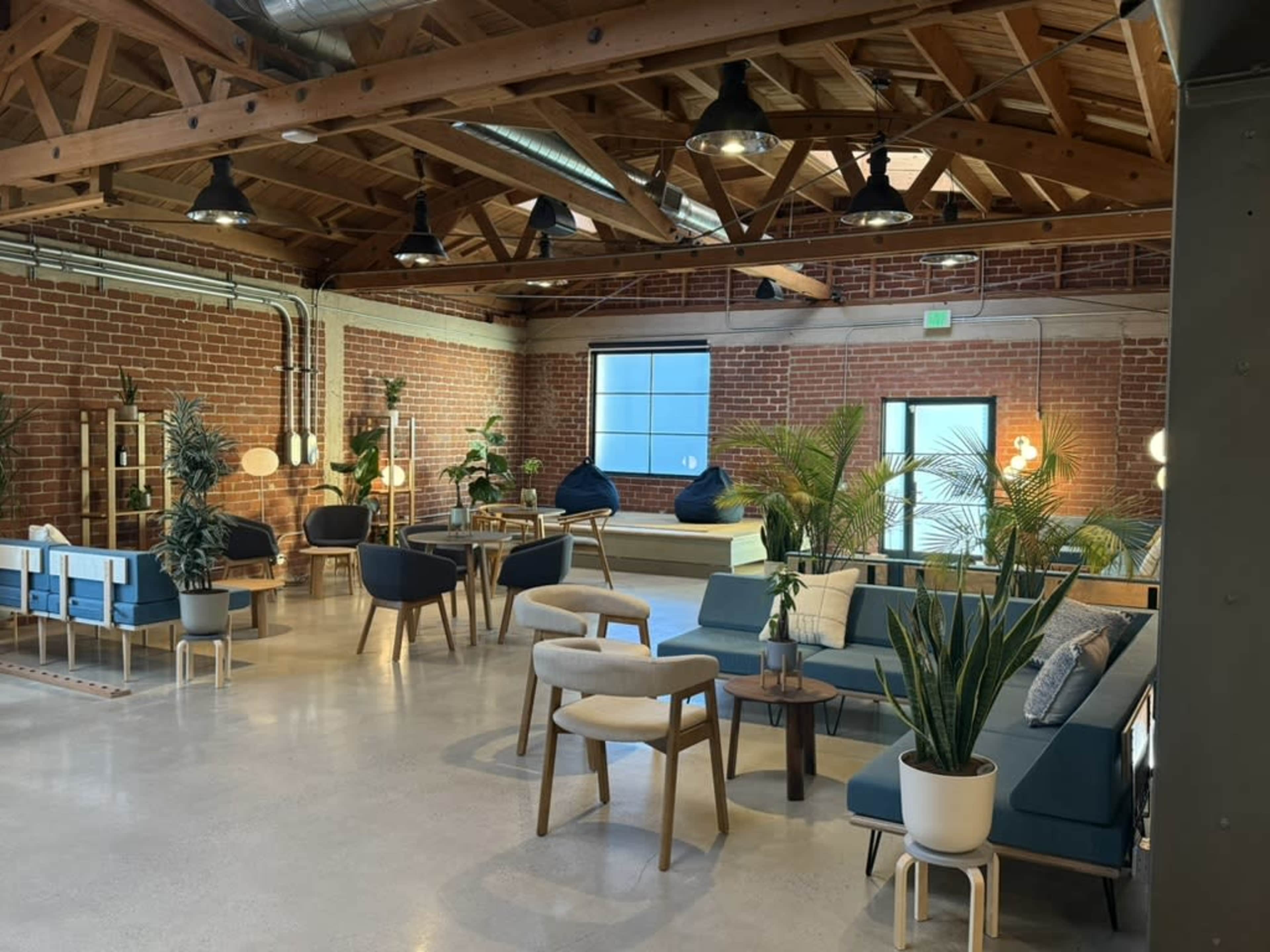 The image shows a spacious, modern lounge area featuring brick walls, wooden beams, and various seating options surrounded by indoor plants.