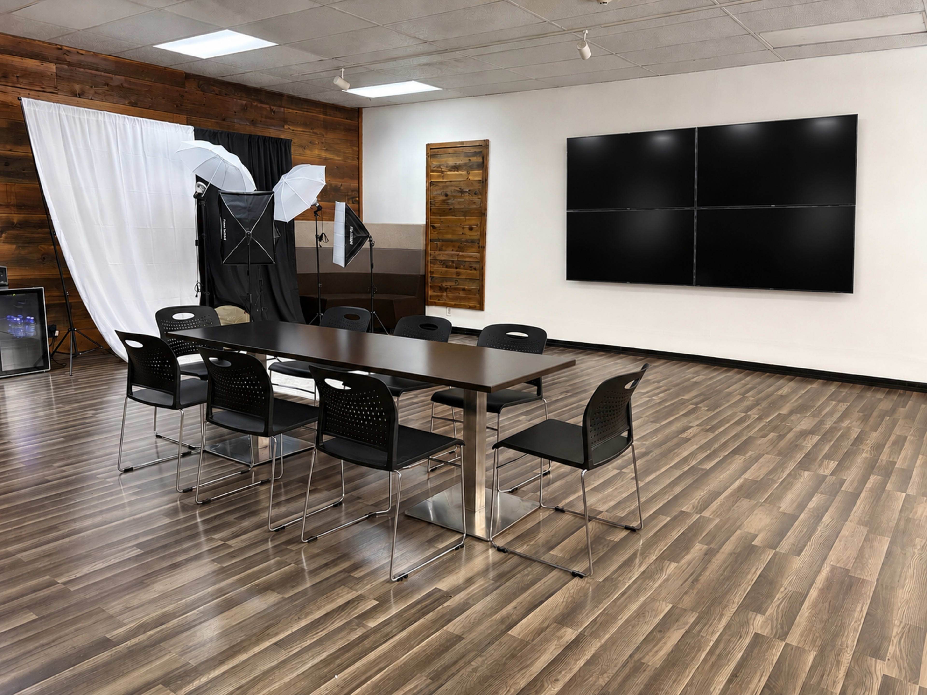 A conference room is set up with a long table surrounded by black chairs, and multiple screens mounted on the wall, with photographic equipment in the background.